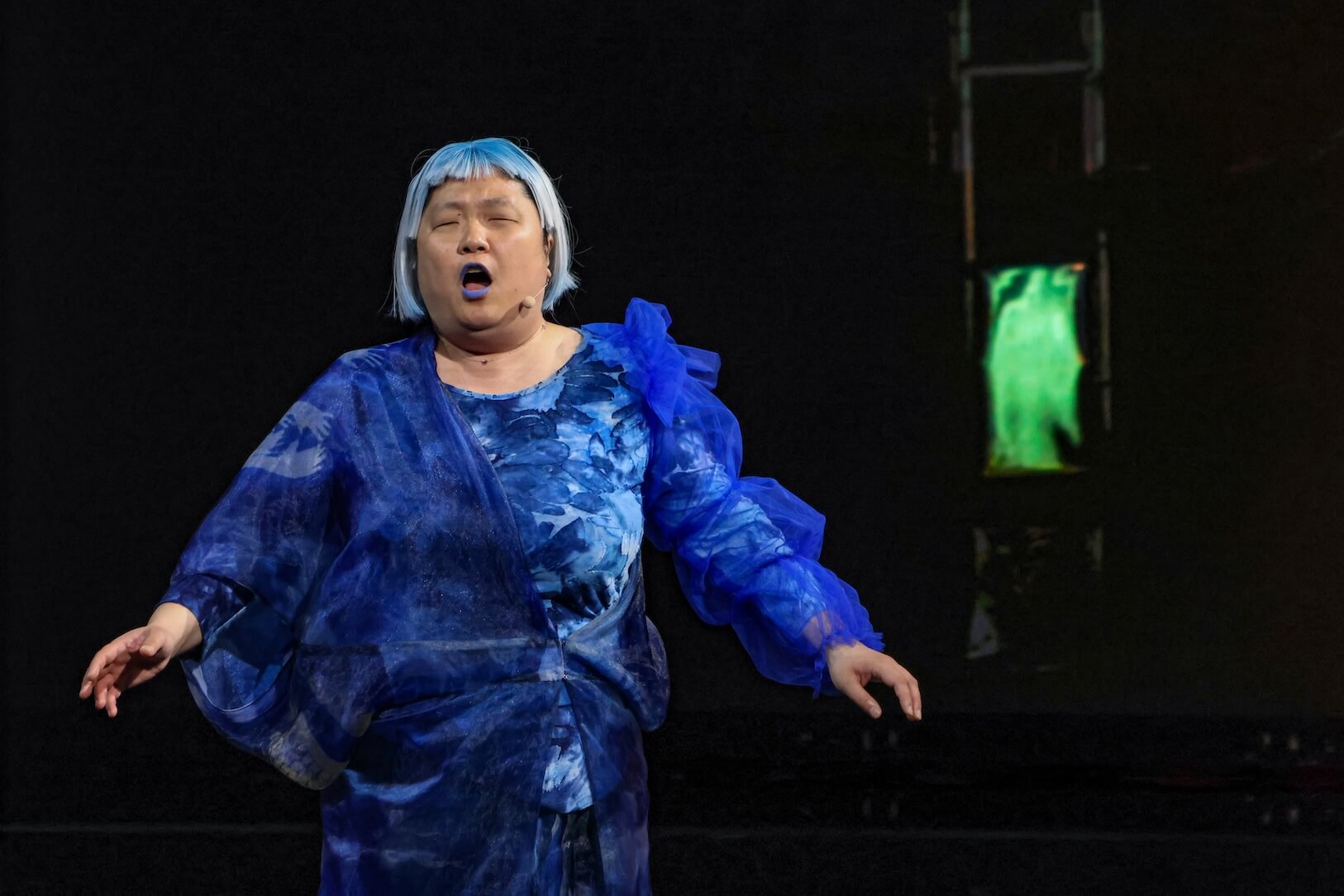 Asian woman performing on stage. She has light blue hair, wearing a flowing, layered blue outfit with various textures. She is singing. Her eyes are closed, as she is Blind. The background is dark, with a faint green light behind them.