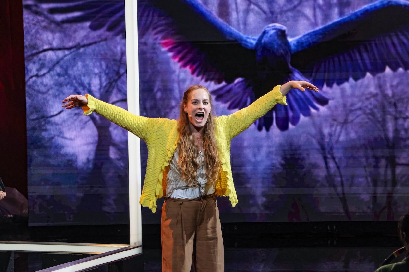 A young woman with long wavy blonde hair standing on stage, arms outstretched like wings. She is wearing a textured yellow cardigan, a light blouse, and brown pants. She is singing. Behind her, a large digital image of a dark blue bird with outstretched wings fills the background.