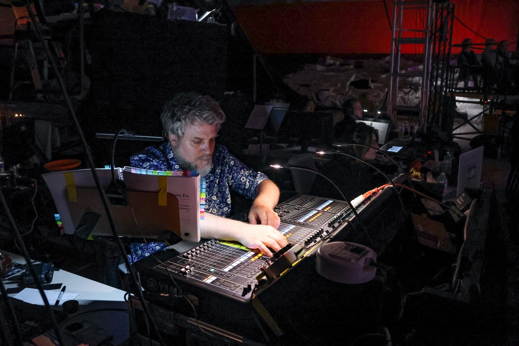 A white man with short curly gray hair and beard, wearing a blue top, working behind the scenes on technical equipment. Others are in the area with him.