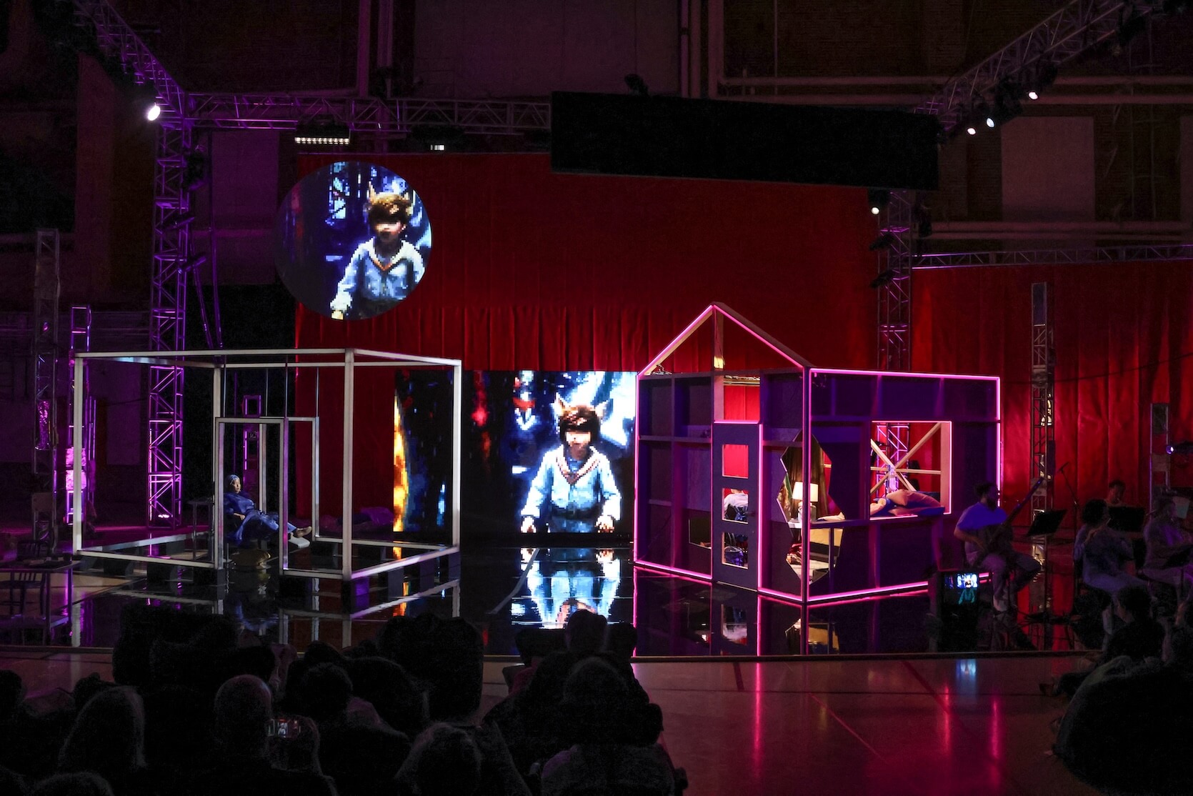 In the center, there are two geometric, house-like structures made from illuminated metal frames. The structure on the left is a simple cubic skeleton, inside which a person dressed in blue is sitting on the floor. The structure on the right is more elaborate, with colored panels forming the outline of a room and a roof, lit by pink-purple light; a person can be seen lounging inside near a small lamp. Behind these structures is a large video screen displaying a child in a blue outfit with fox ears. Above this, a circular video screen projects the same or similar image of the child. The background is draped with red fabric. The lighting throughout is dramatic, with purple and pink tones dominating, and some members of an orchestra or band can be dimly seen playing at the right edge. The audience is in the foreground.