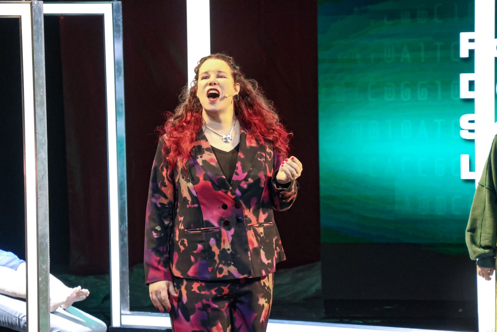 A woman with long curly brown and red hair wearing a vibrant, patterned suit in shades of pink, red, and black, standing on a stage. She is mid-performance, singing, with her mouth open and a dramatic expression. She is wearing a necklace and has a microphone headset on. The background features illuminated vertical frames and a teal-blue screen with abstract text and design, creating a modern, theatrical atmosphere. A person's legs can also be seen in the left part of the image, lying on a bed.