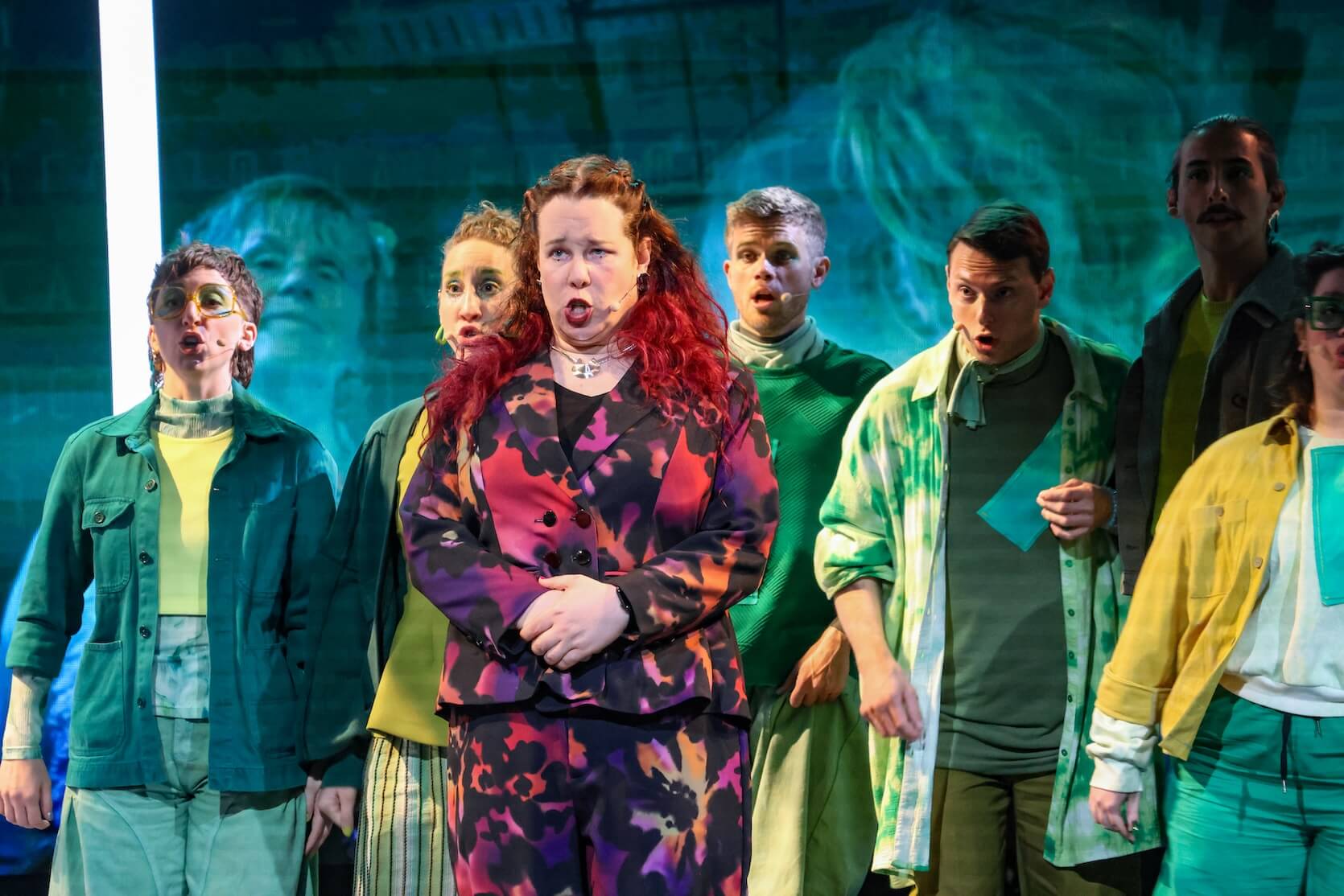 A stage scene with a group of people standing close together, singing or speaking with expressive faces. The person in the center front has long reddish hair and is wearing a colorful suit with a purple and orange pattern, while others around them are dressed in shades of green, yellow, and teal, with some in patterned clothing. There is a blue-toned background with a projected image.