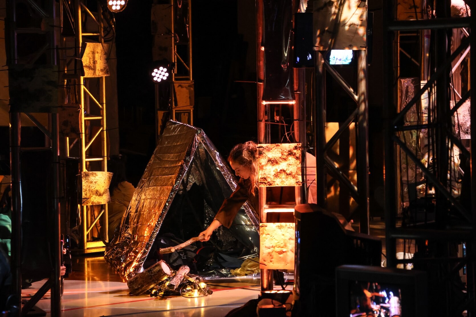 A person crouching in front of a small, triangular reflective tent, feeding faux logs into a campfire setup made of electric lights and wood pieces. The scene is staged within a structure of tall metallic trusses, illuminated by various warm, theatrical lights and decorated panels, suggesting an indoor performance or art installation with a camping theme. The environment is busy with props and layered lighting.