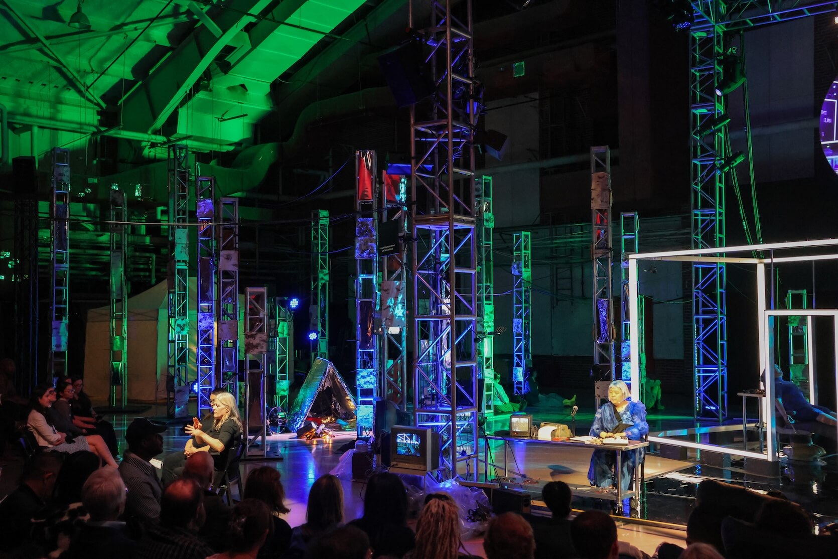 Theater stage with colorful lighting. The image shows a large, industrial-looking stage setup, filled with metal scaffolding structures lit in green, blue, and purple hues. There are two main performers: one person sits at a small table on the right side of the stage, dressed in a blue outfit, appearing to be engaged in reading or speaking. Another person sits on a chair closer to the audience on the left, gesturing expressively, possibly interpreting or narrating. The audience is seated closely in rows, attentively watching the performance. There are various objects and screens scattered on stage.