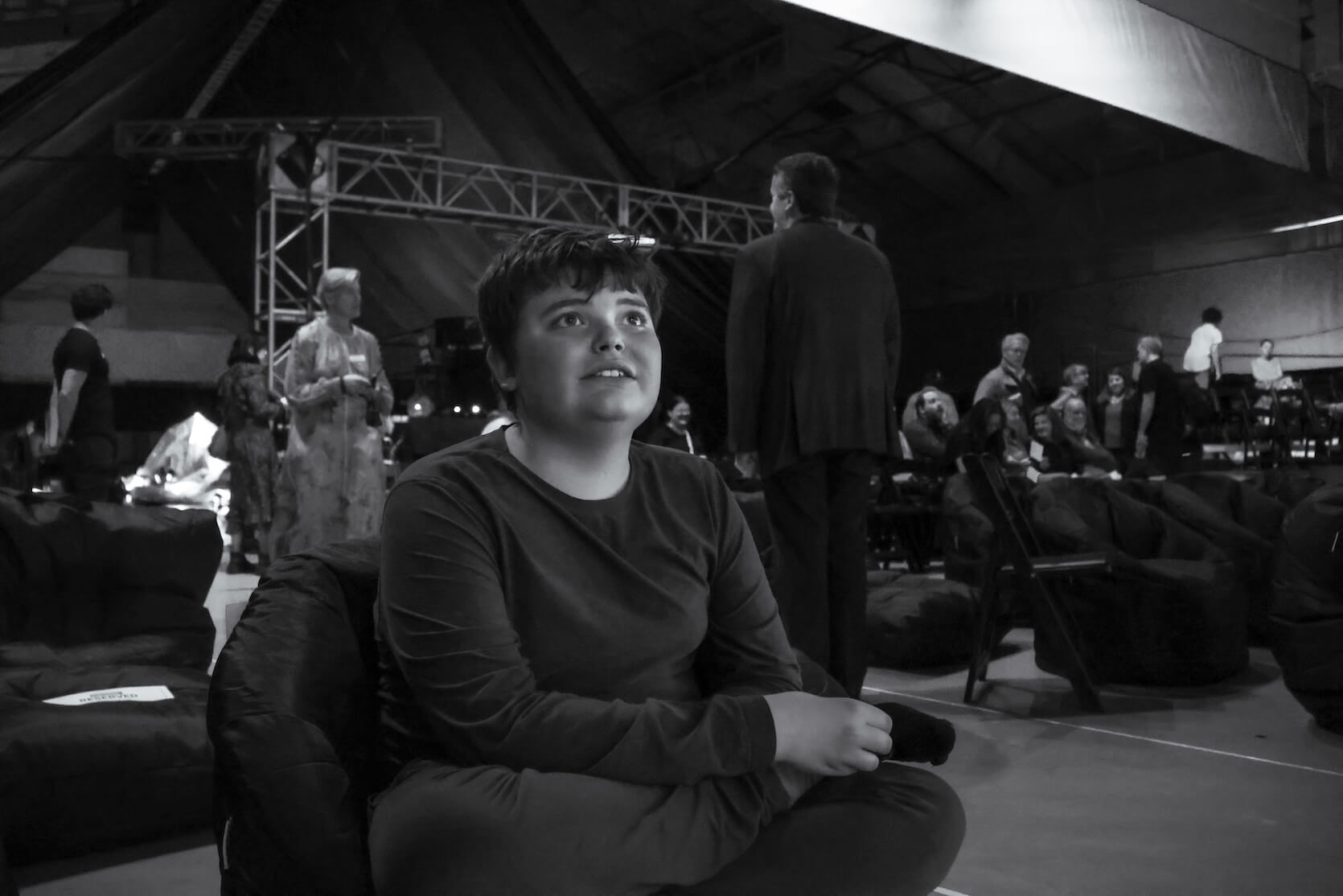 A young boy sitting cross-legged in a beanbag chair. The image is in black and white. The boy is looking slightly upward and smiling softly, wearing a long-sleeved shirt and pants. In the background, there are several people standing, sitting, and talking, surrounded by additional beanbag chairs and folding chairs.