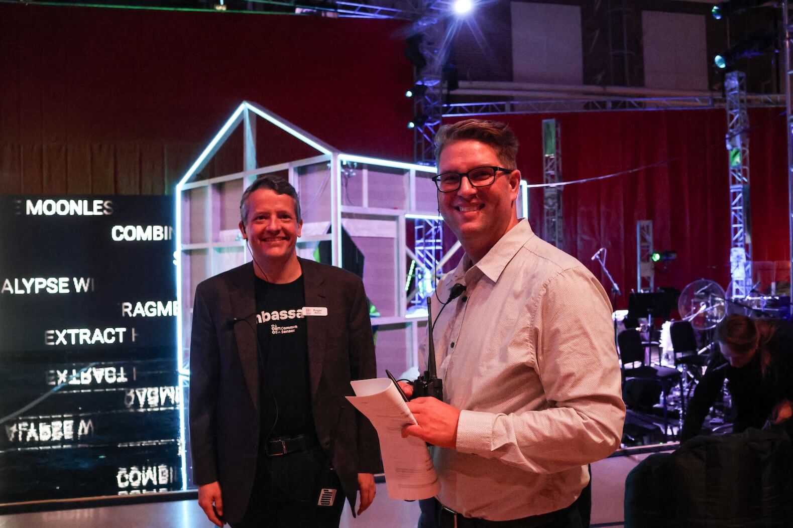 Two men standing together, smiling. The left one is a white man with short hair, wearing an all black outfit: shirt, blazer, pants. The other on the right is a white man with short brown hair, wearing glasses and a light button up shirt. They are holding/wearing tech equipment (walking talkies, etc.) They are standing in front of a stage set.