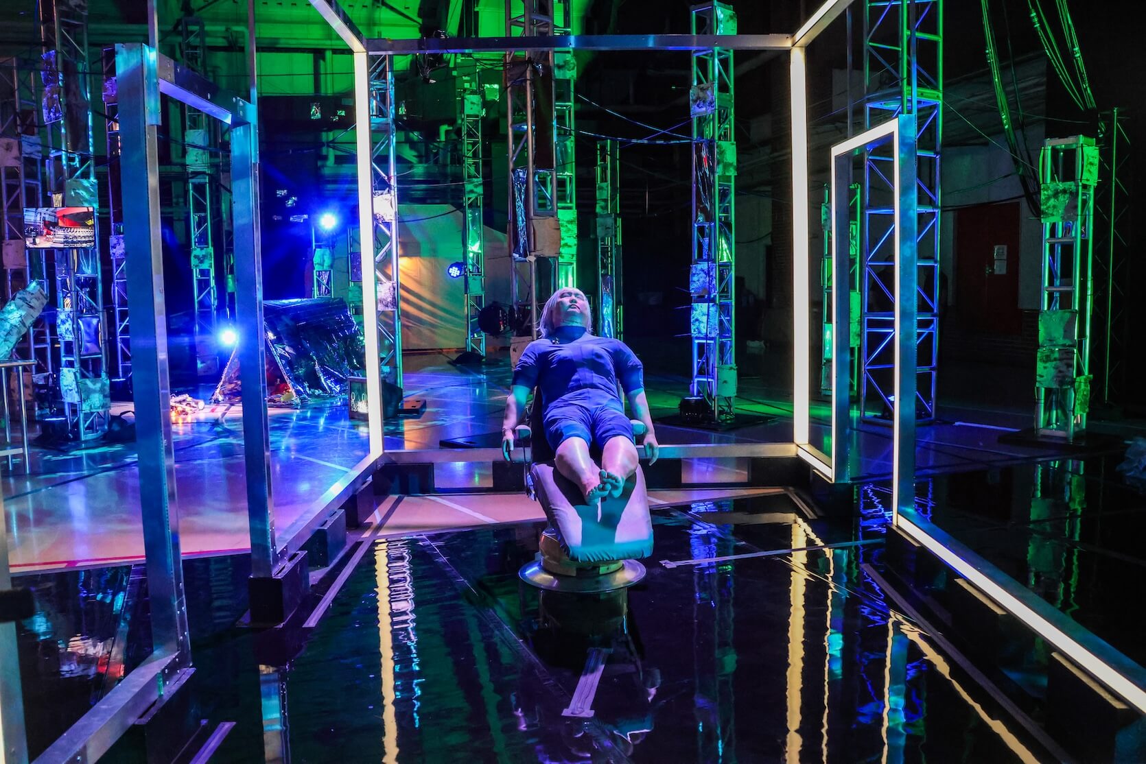 A human-shaped crash test dummy in a blue suit strapped to a reclining chair, positioned in the center of a futuristic testing area, illuminated by vibrant blue, green, and purple lights, creating strong reflections on the shiny black floor. The testing area is framed by large square metal structures, and metallic structures with attached electronic equipment and lights line the background.
