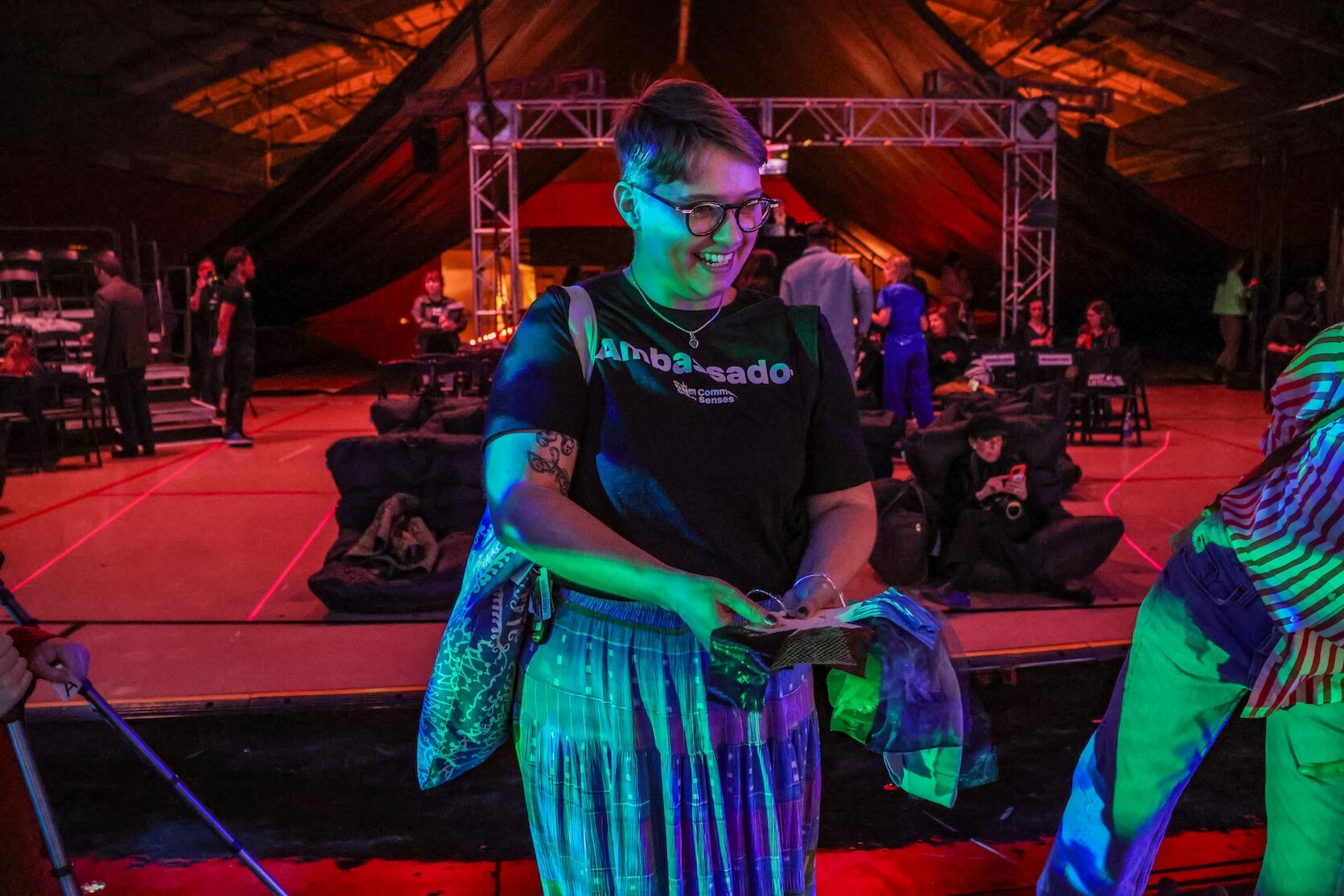 A smiling person wearing glasses and a black t-shirt that says "Ambassador,” standing indoors at an event or party with colorful lighting. They have tattoos, short hair, a tattoo on their right arm, and are holding items in their hands. They are also carrying a patterned bag and wearing a pleated, blue-and-green skirt. The background features bean bags, people sitting and standing, some equipment, and a structure with lights, all under a dramatic, tent-like ceiling illuminated in warm orange and cool blue tones.