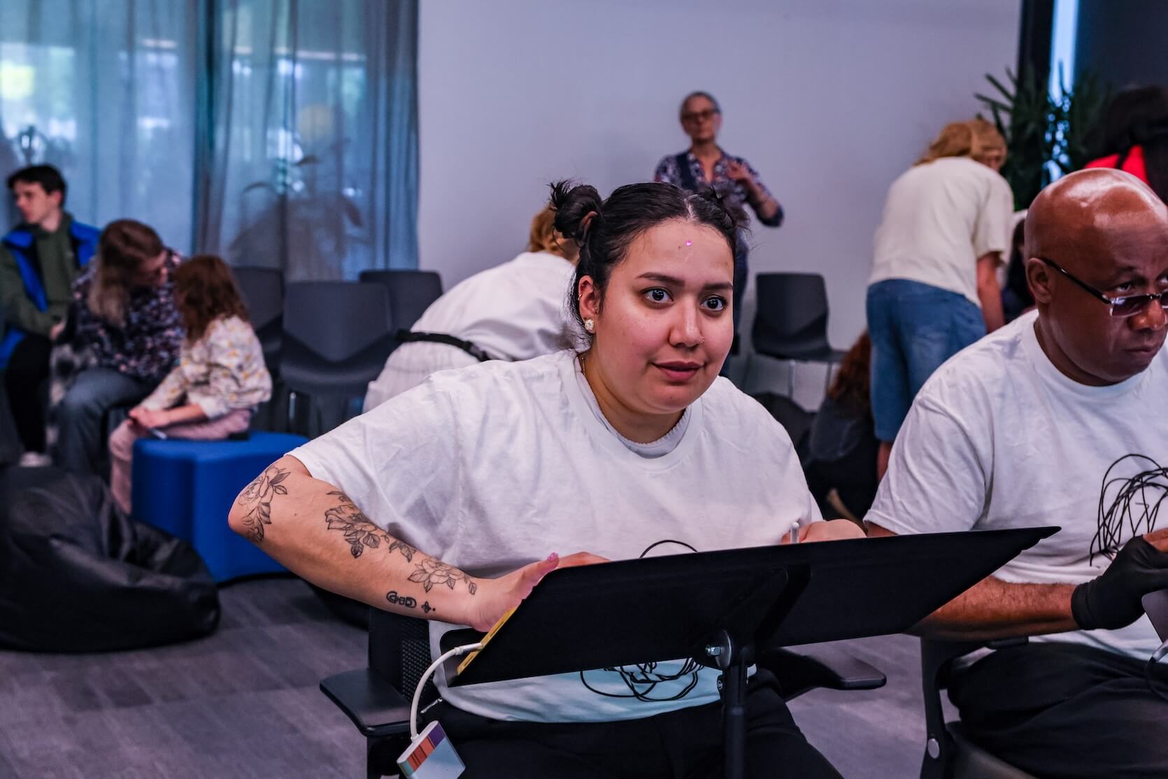 A Latina woman with her hair pulled back in space buns, wearing a white short sleeve t-shirt with scribbles on it. She has tattoos on her arm. She is holding an Apple pencil in one hand, and is focused on what is in front of her. Other people are in the background.