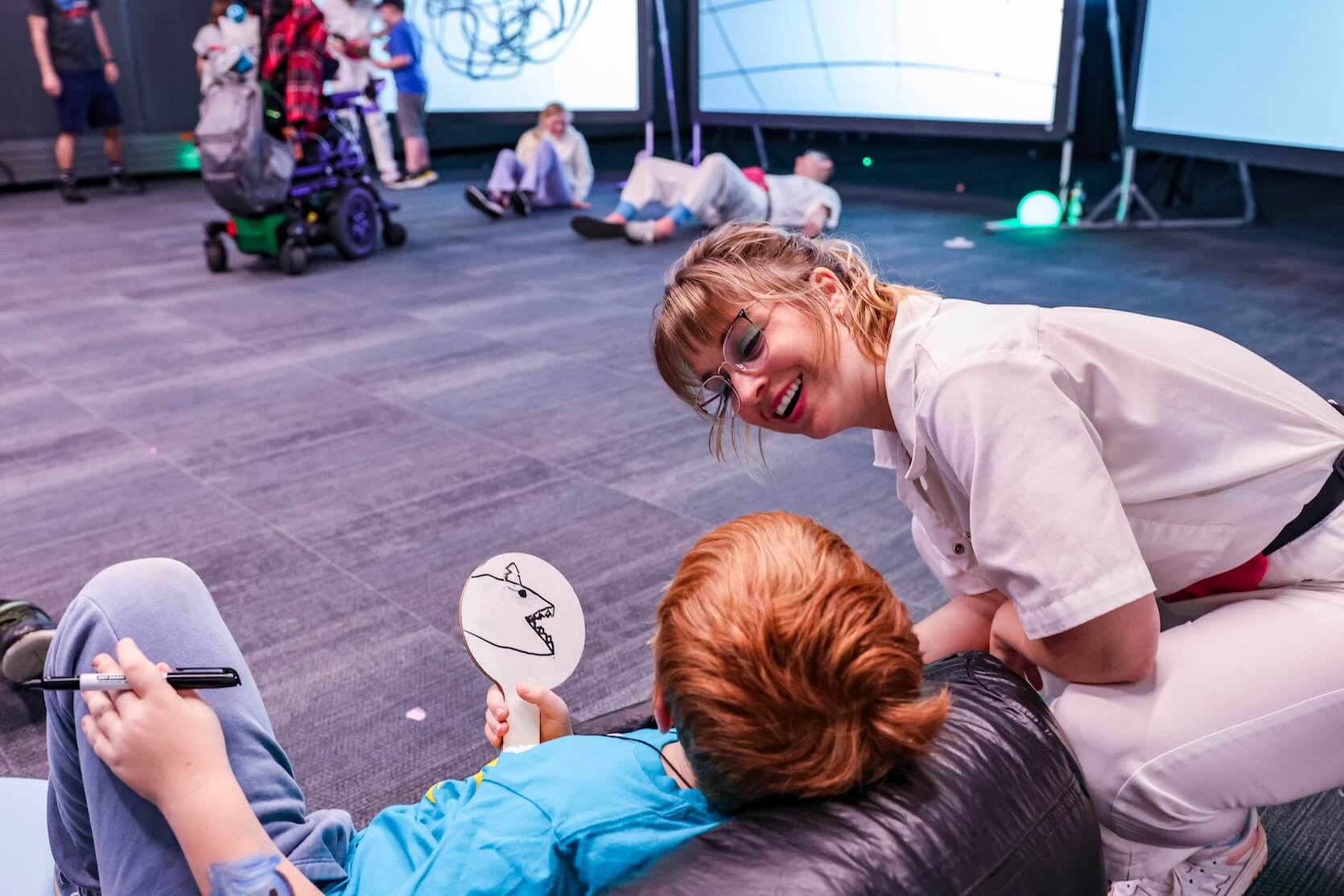 A white woman with blonde hair with bangs pulled up in a ponytail, wearing glasses and a white jumpsuit, smiling at a young redheaded boy who is laying on a bean bag. There are other people in the background.