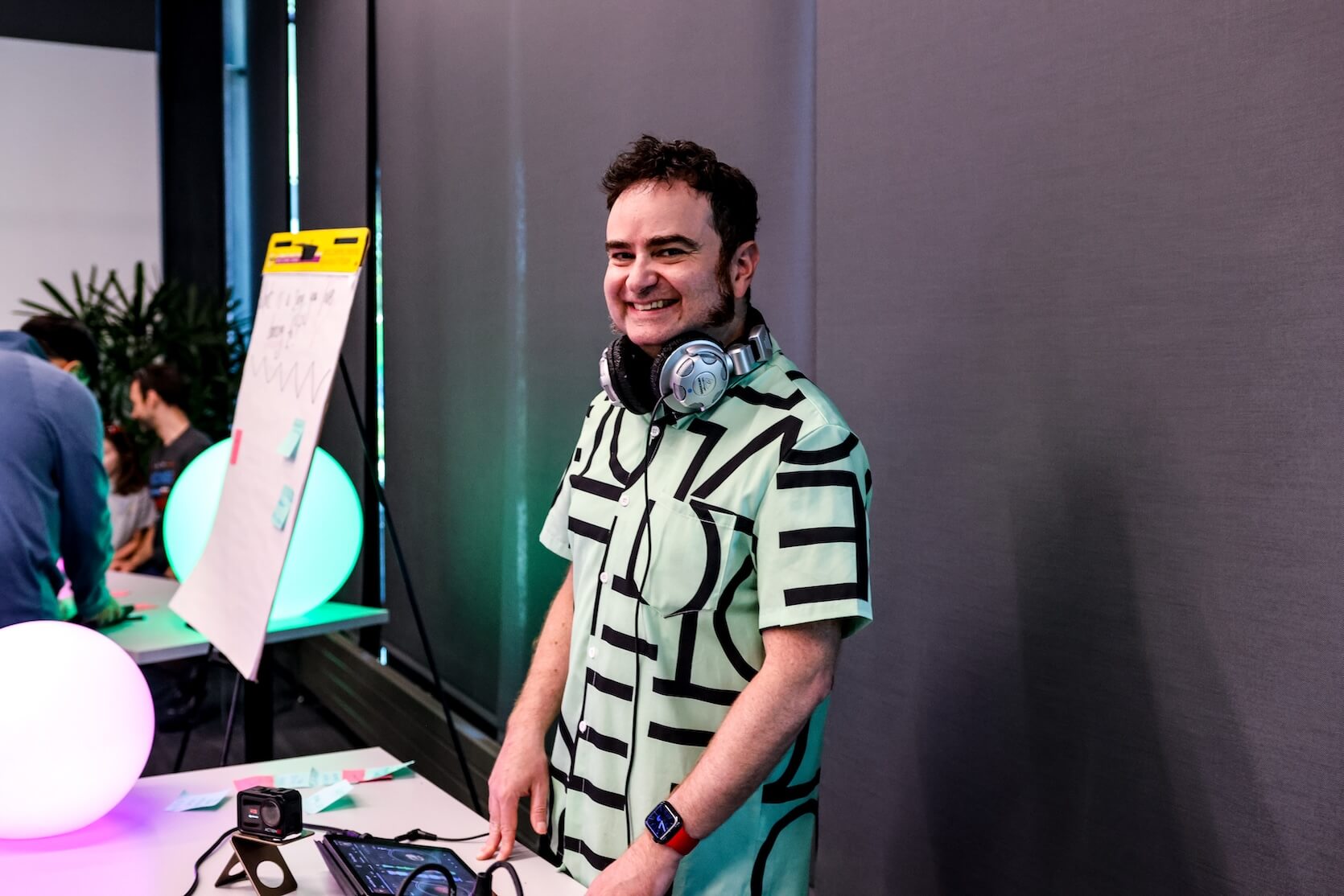 A white man with short brown hair and sideburns, wearing a graphic button up shirt, headphones around his neck, smiling. He is the DJ.