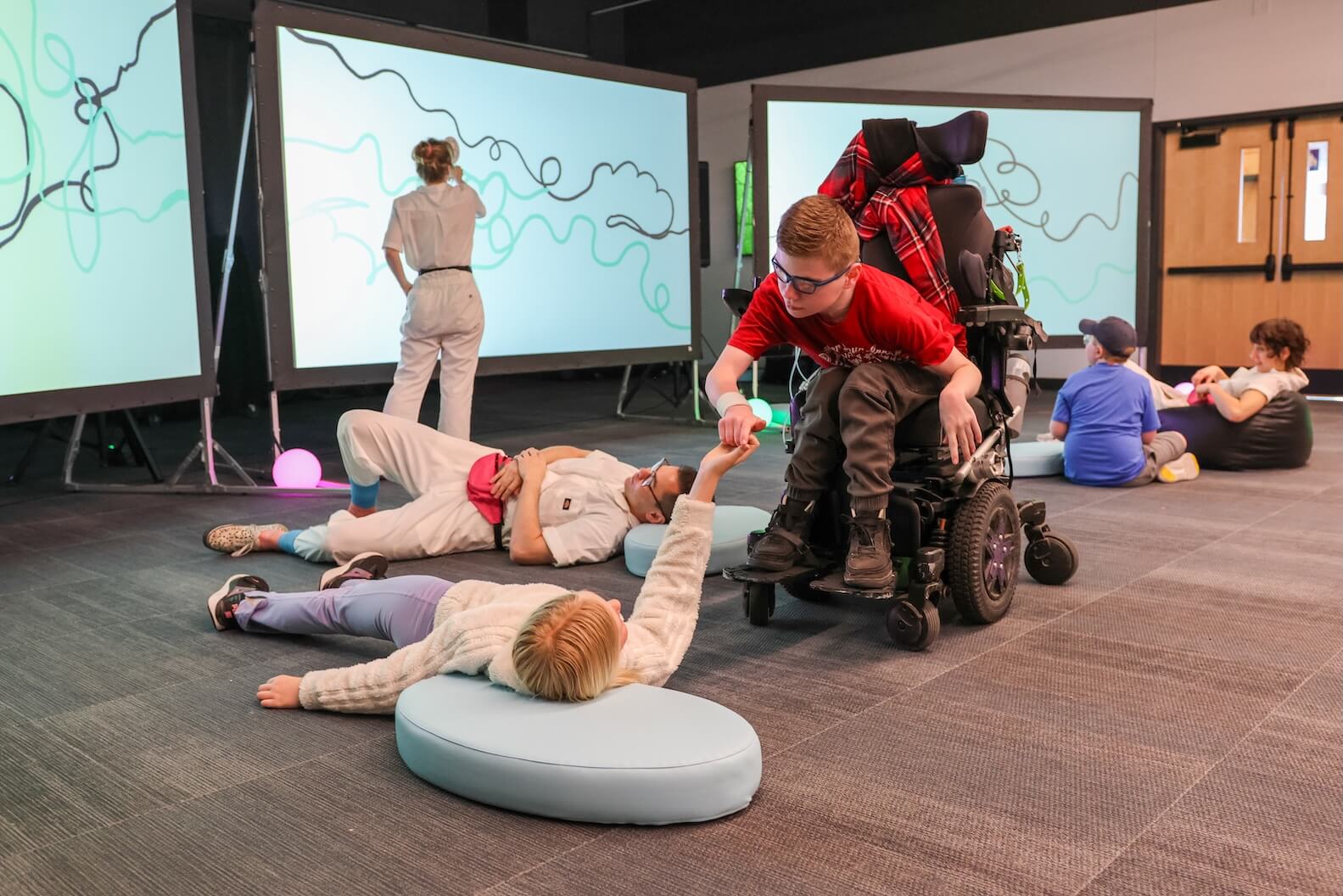 A young boy with short red hair, glasses, sits in a powerchair, holds the hand of an attendee who is laying on the floor. Four other people are in the background, one standing and three also laying on the ground.