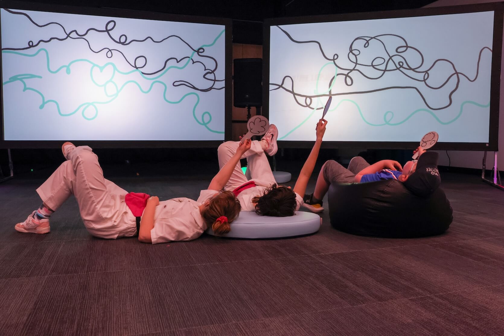 Three people lay on the floor, holding up paddles. The two on the left are in Risky Disco white jumpsuit uniforms. The other is an attendee.