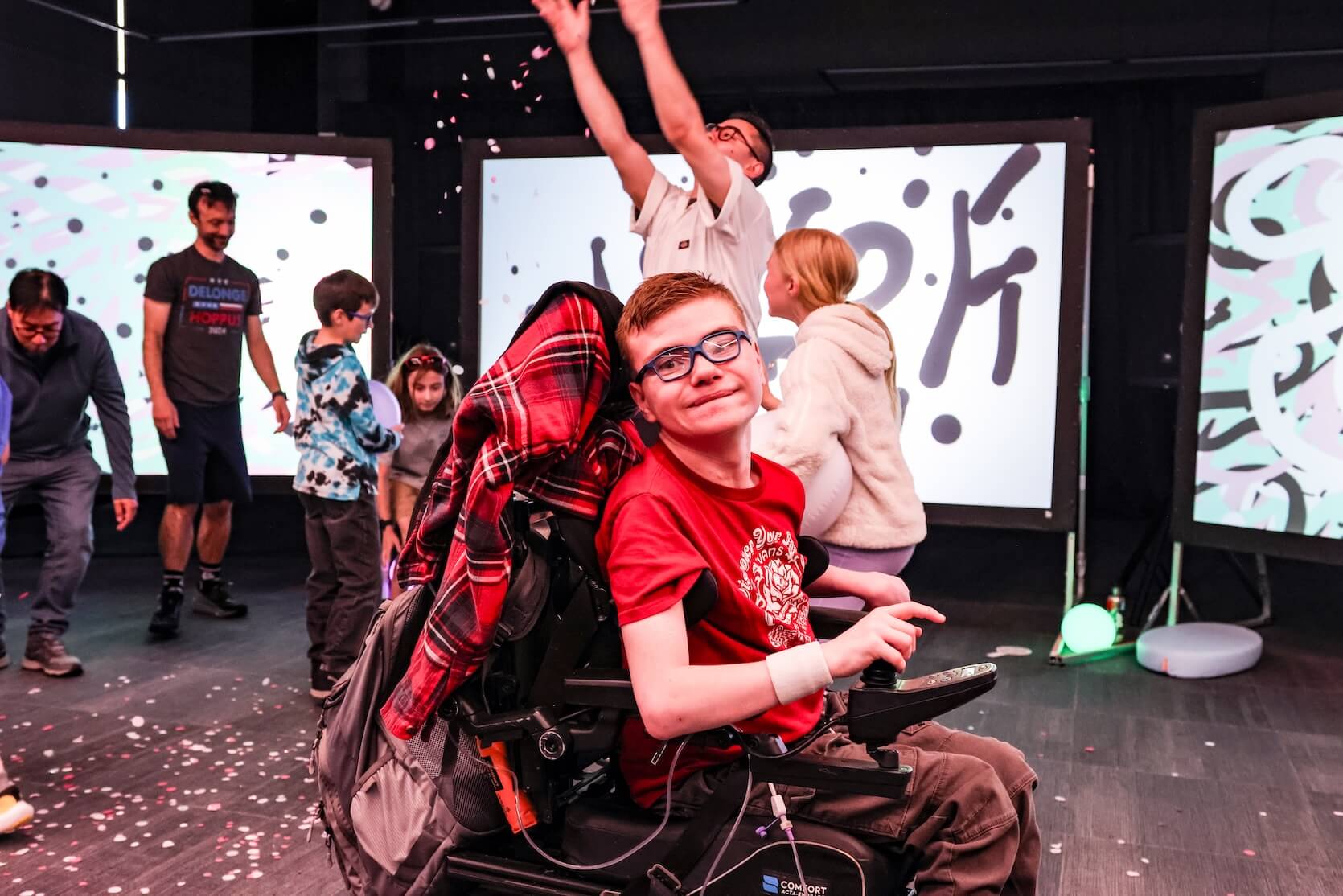 A young boy with short red hair, glasses, sits in a powerchair, smiling. He is wearing a red graphic tee and dark cargo pants. A red flannel jacket sits on his powerchair. There are other people in the background.