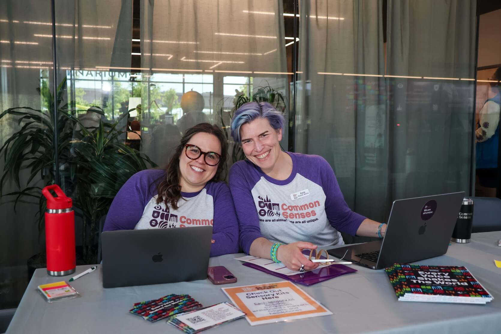 Two white women sitting together at a table, smiling. The woman on the left has long brown hair, half pulled back. She is wearing glasses and a Common Senses jersey tee. The woman on the right has short purple and blue hair, and is also wearing a Common Senses jersey tee. Laptops are open in front of them. Marketing materials are laid out on the table.