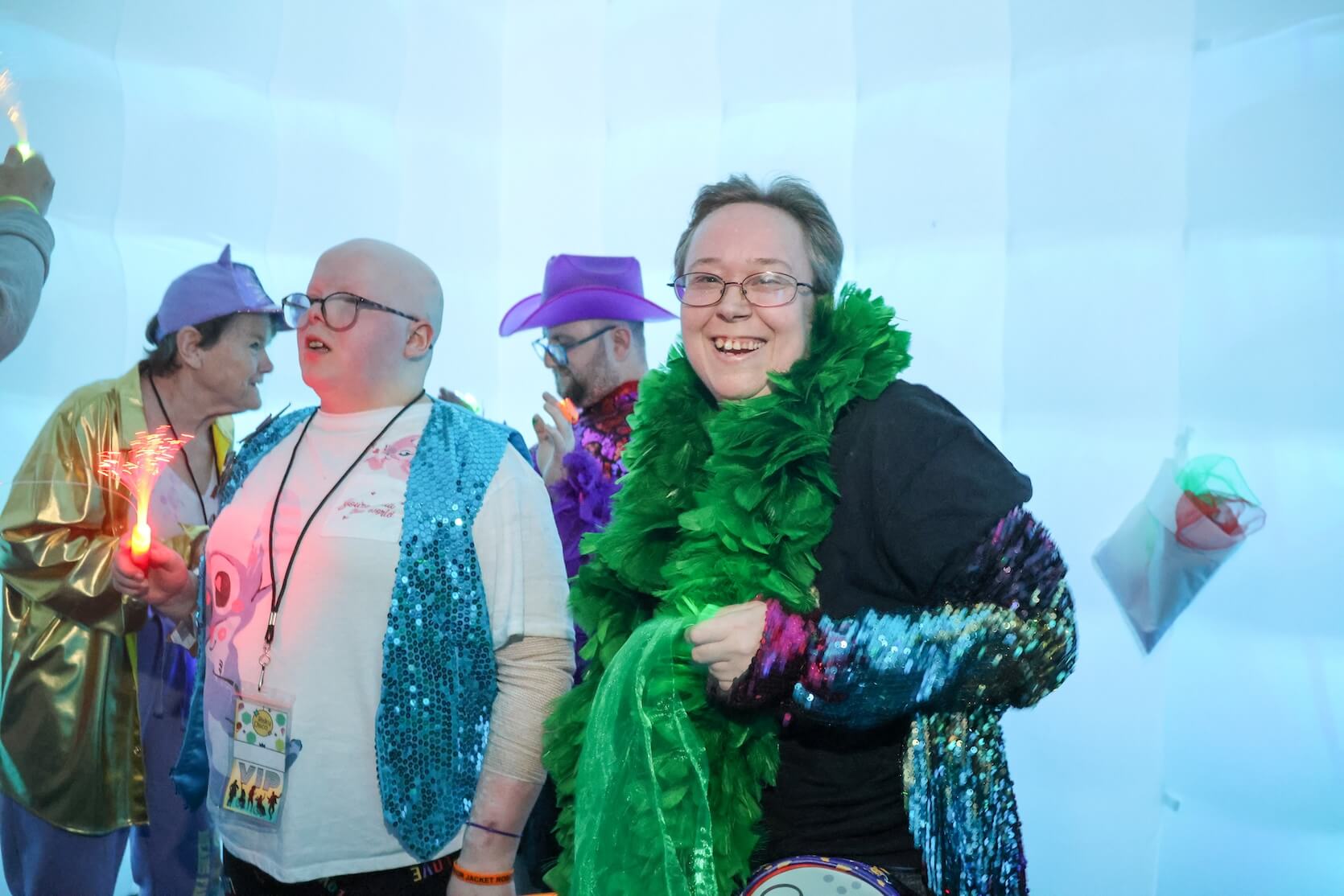 A white person with short hair, wearing glasses, a green feather boa, a black shirt on top of multicolor sequin attire, giving a big smile at the camera. There are other people in the background.