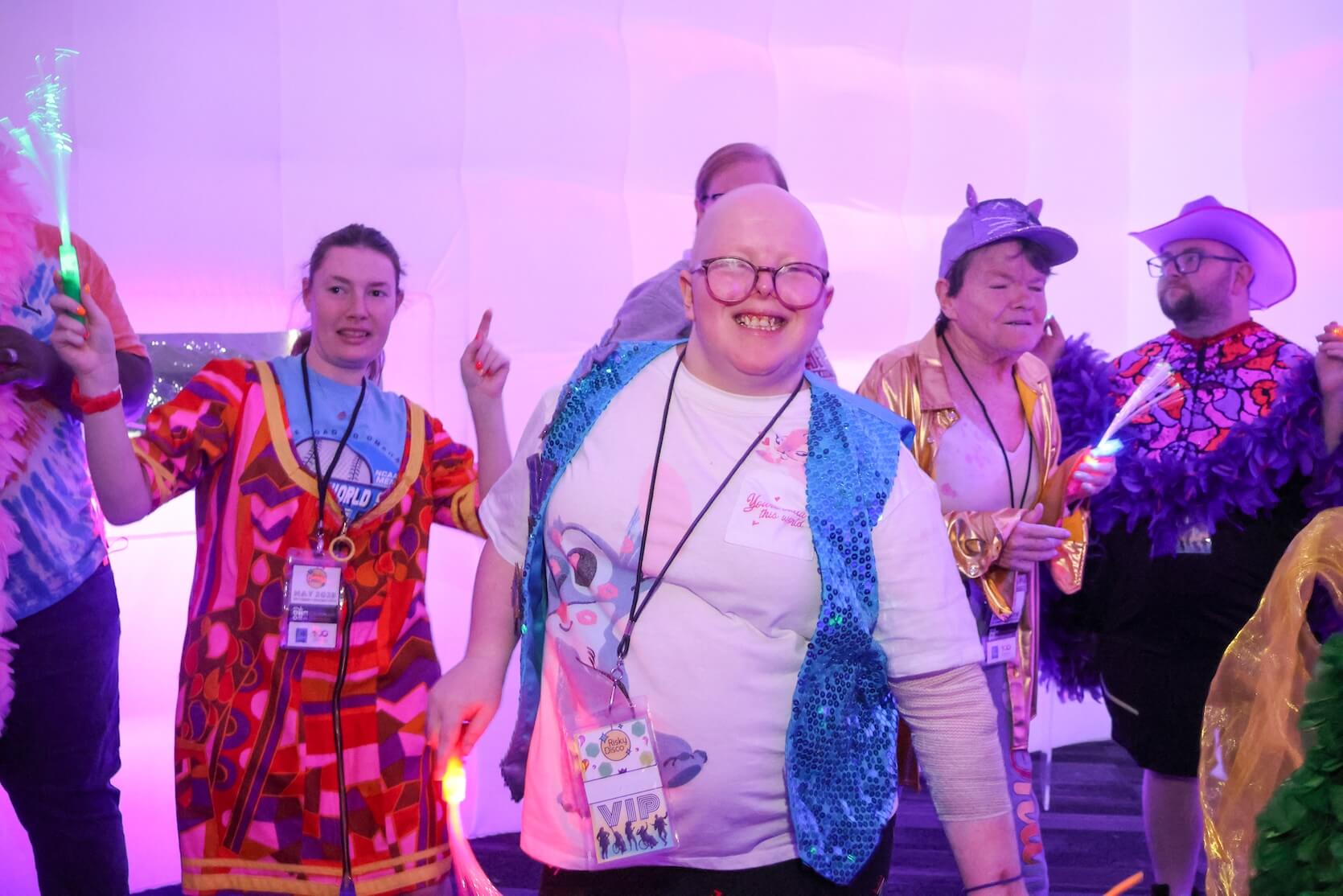 Focus on a bald white person wearing glasses, a Stitch top with a blue sequin vest on top, with a large smile. Other people are in the background behind them.