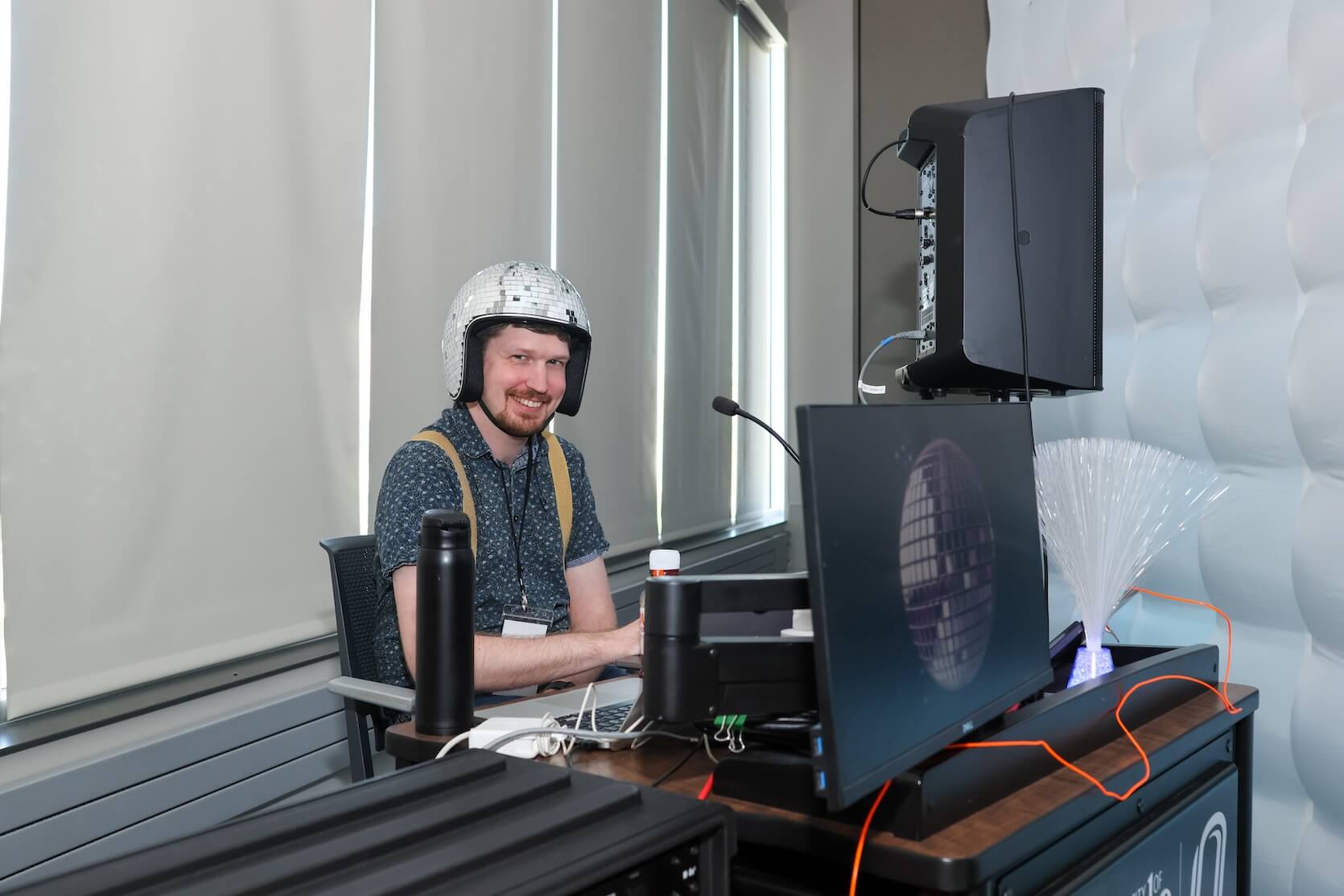A white man with short brown hair, wearing a disco helmet, a blue top, and carrying a backpack, sits and smiles at the camera. He is in front of a laptop and speaker.