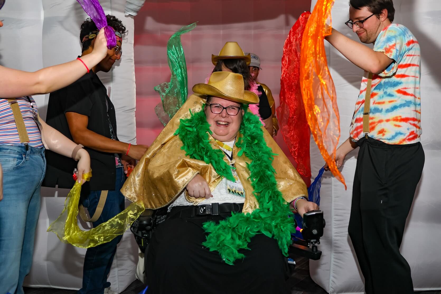 A person in a wheelchair, wearing a long sleeve shirt, jeans, and a green cowboy hat. They are surrounded by four people holding scarves, presumably dancing with them around said person.