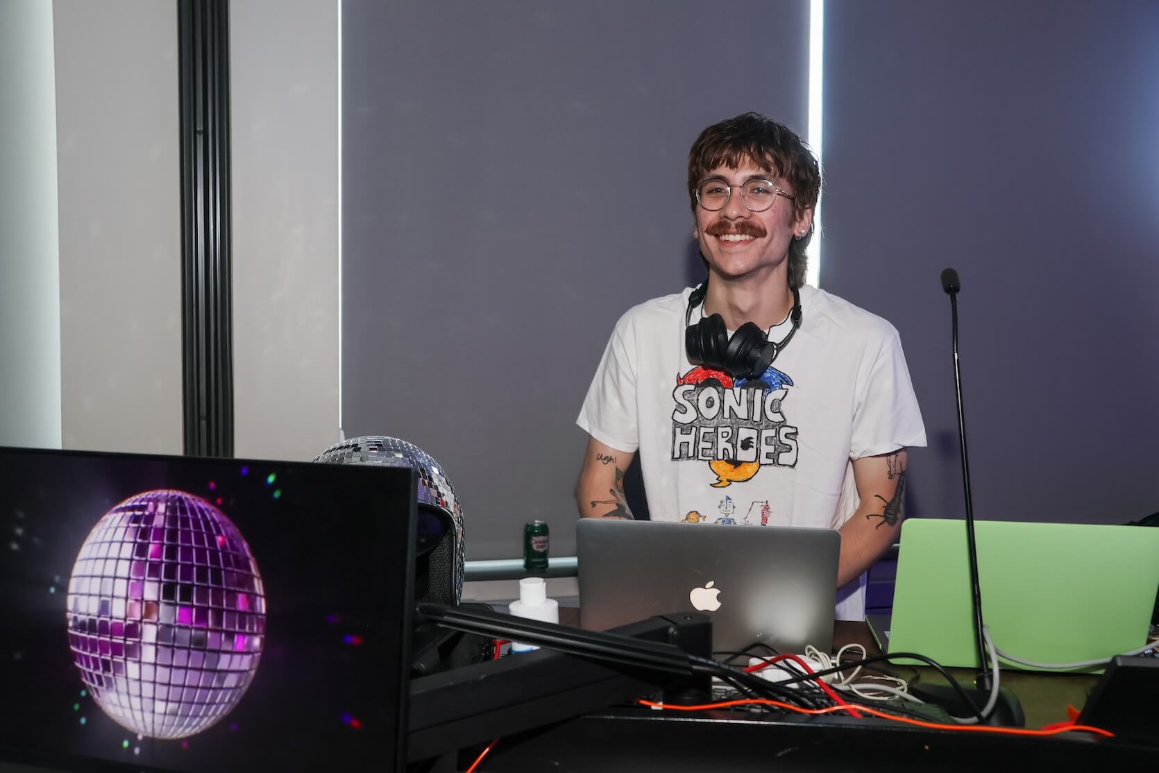 A young white man with brown hair and a mustache, wearing glasses and a Sonic Heroes t-shirt. There are tattoos on his arm and a pair of headphones around his neck. He is the DJ.