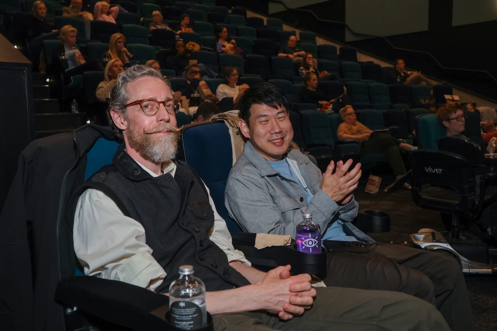 A white man has short brown and gray hair with a beard and mustache, wearing a long sleeve white shirt with a black vest. The other is an Asian man with short black hair, wearing a gray shirt and light brown-gray jacket. They are sitting beside each other at the theater, smiling at the camera. Other audience members join them in the background.
