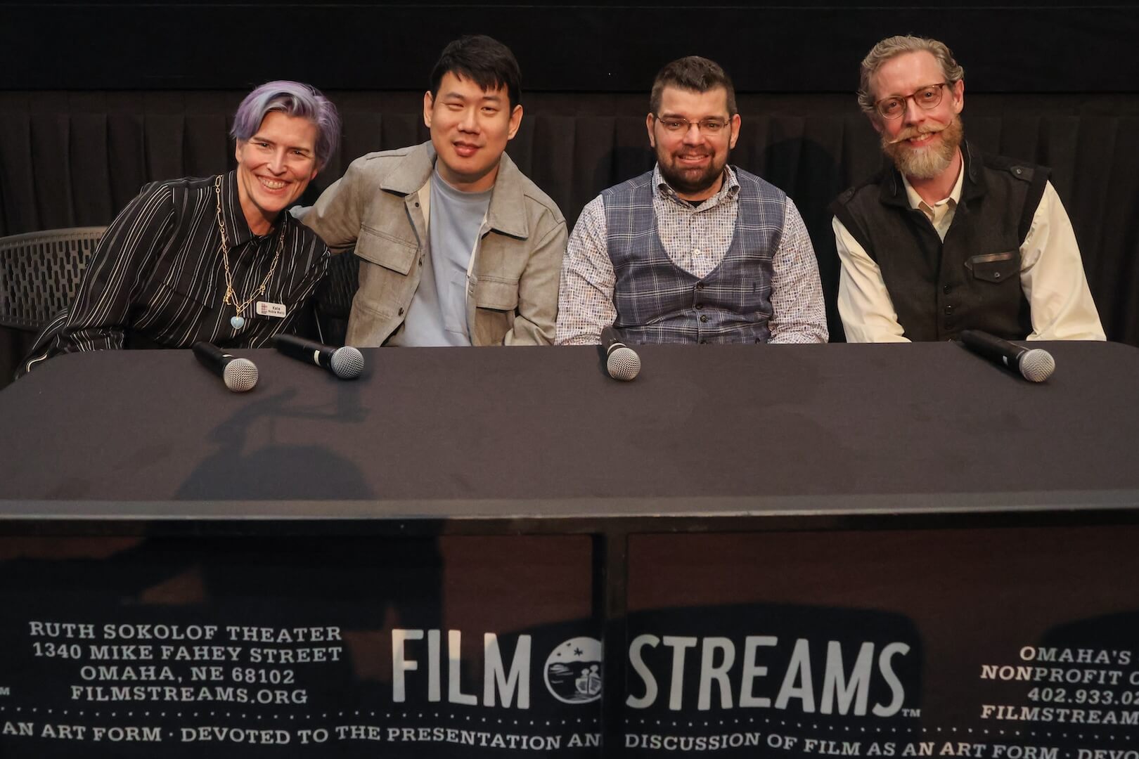 Four people sitting together at the front of the theatre, smiling: a white woman has short blue and purple hair, wearing a white and black striped dress and jewelry; an Asian man has short black hair, wearing a gray shirt and light brown-gray jacket; a white man has short brown hair and beard, wearing glasses and a textured graphic button up with a blue vest; and the last white man has short brown and gray hair with a beard and mustache, wearing a long sleeve white shirt with a black vest.
