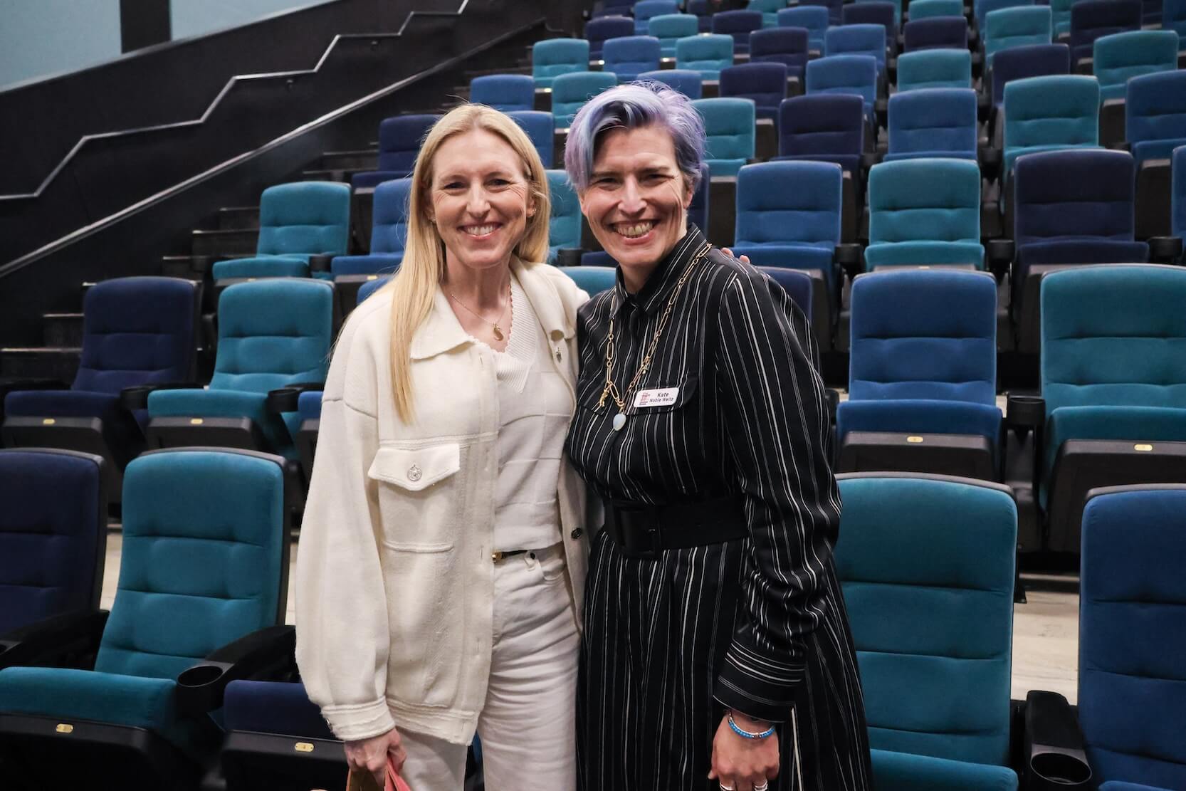 Two white women stand together, smiling. The woman on the left has long blonde hair, and is wearing all white: shirt, pants, and jacket. The woman on the right has short blue and purple hair, wearing a white and black striped dress and jewelry. They are standing in front of many chairs in various shades of blue.