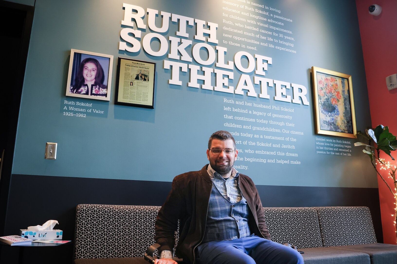 A white man with short brown hair and beard, glasses, and wearing business casual attire sits in a chair, smiling. He is sitting in front of a wall that says Ruth Sokolof Theater, where there is a framed photo of Ruth herself.