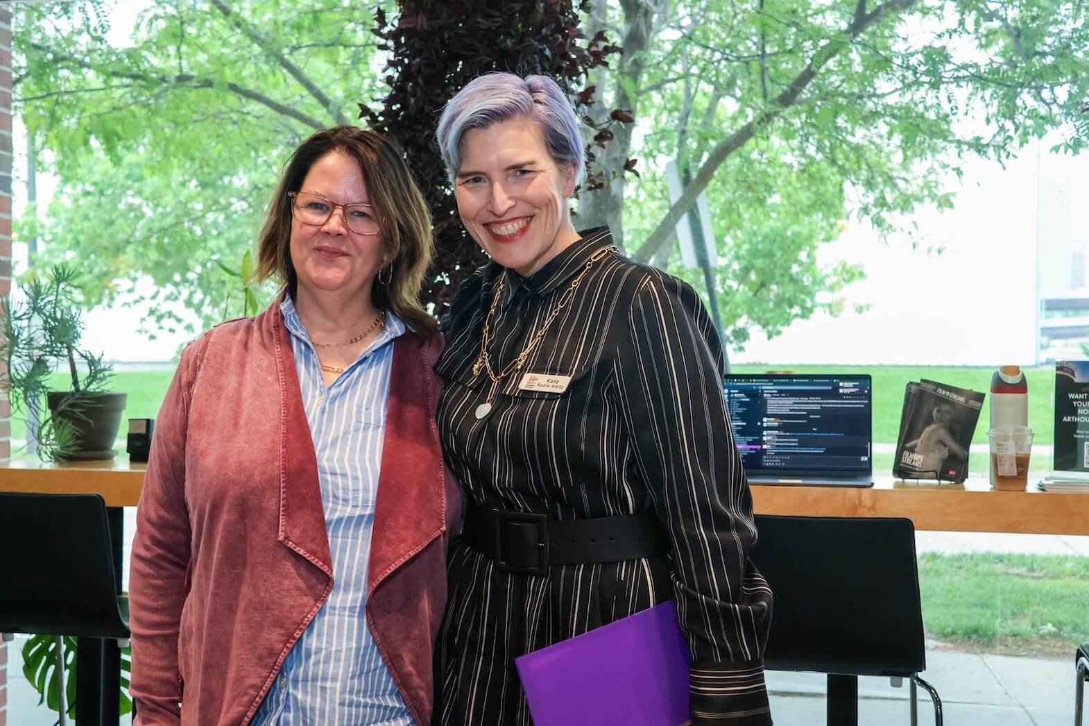 Two white women stand together, smiling. The woman on the left has shoulder length brown hair. She is wearing glasses, a striped blue and white button top, and a light red blazer/jacket over it. The woman on the right has short blue and purple hair, wearing a white and black striped dress and jewelry.