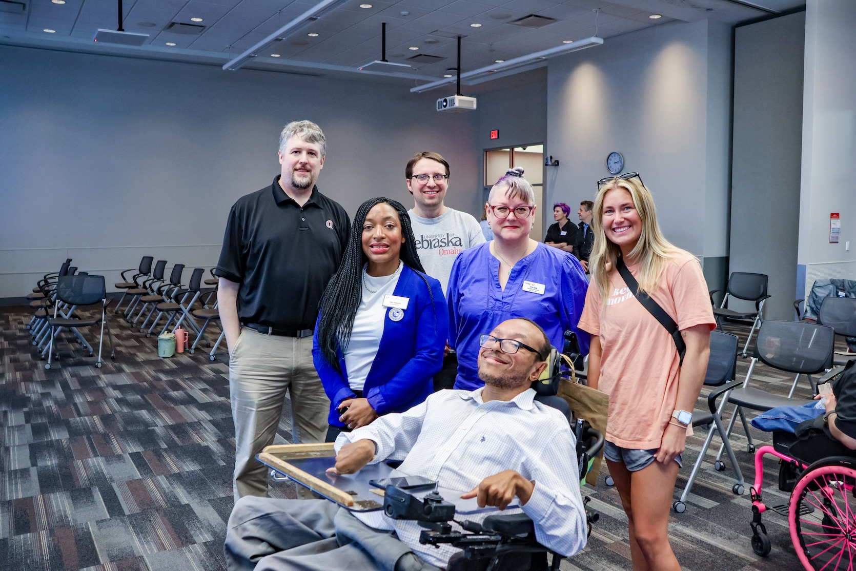 A Black man, a Black woman, and two white men, and two white women pose together for a photo. The Black man sits in a powerchair. The rest are standing.