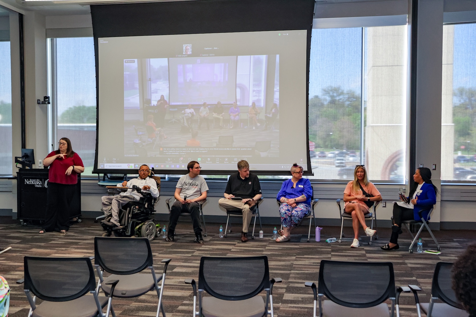 A Black man, a Black woman, and two white men, and two white women sit for a panel. One woman is speaking. The Black man is in a powerchair while the others are in regular chairs. There is an ASL interpreter standing beside them.