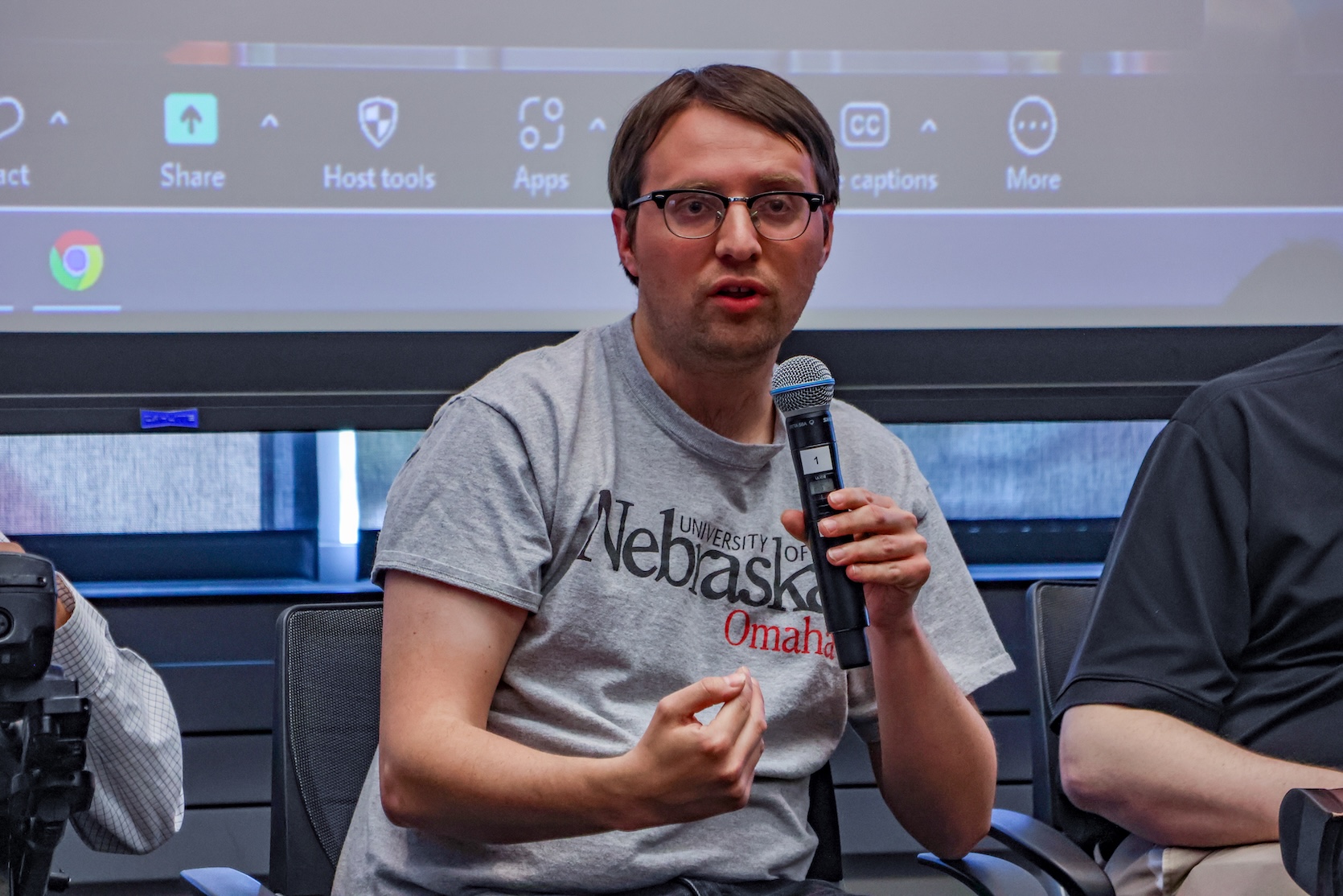 A white man with short brown hair combed over on both sides. He is wearing glasses, a gray UNO shirt. He is holding a microphone and speaking.