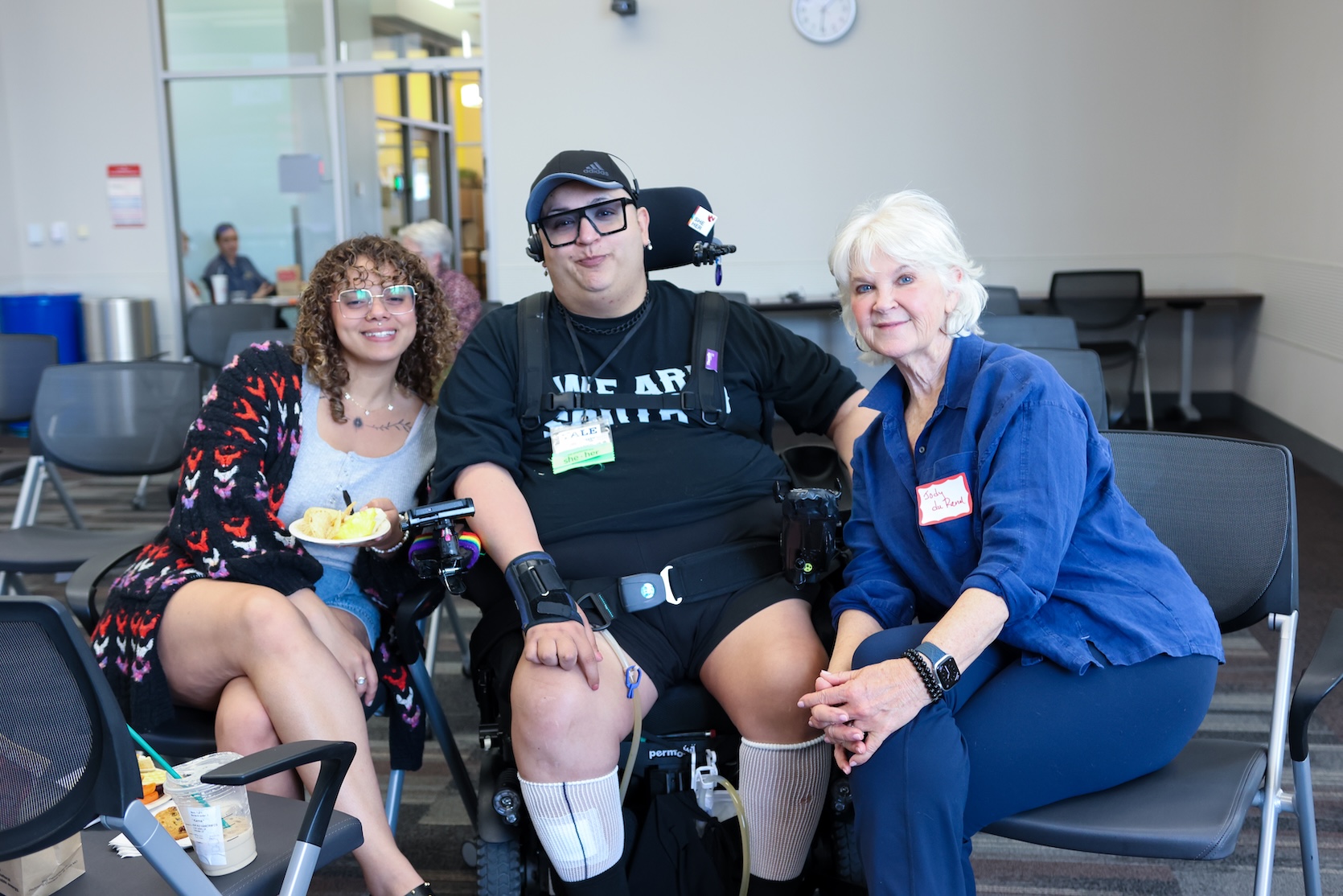 Three people sitting together for the camera. The person in the middle is sitting in a powerchair. The other two on either side of them are sitting in regular chairs.