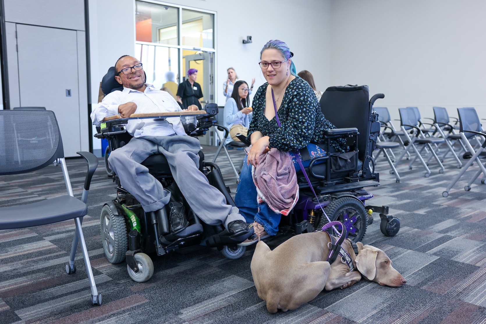 A Black man with glasses, wearing a white gingham button up and gray slacks, sits in his powerchair. A white woman with long blue and purple hair, wearing a floral top and jeans, sits in hers. The woman has a service dog with her. The dog is laying on the floor.