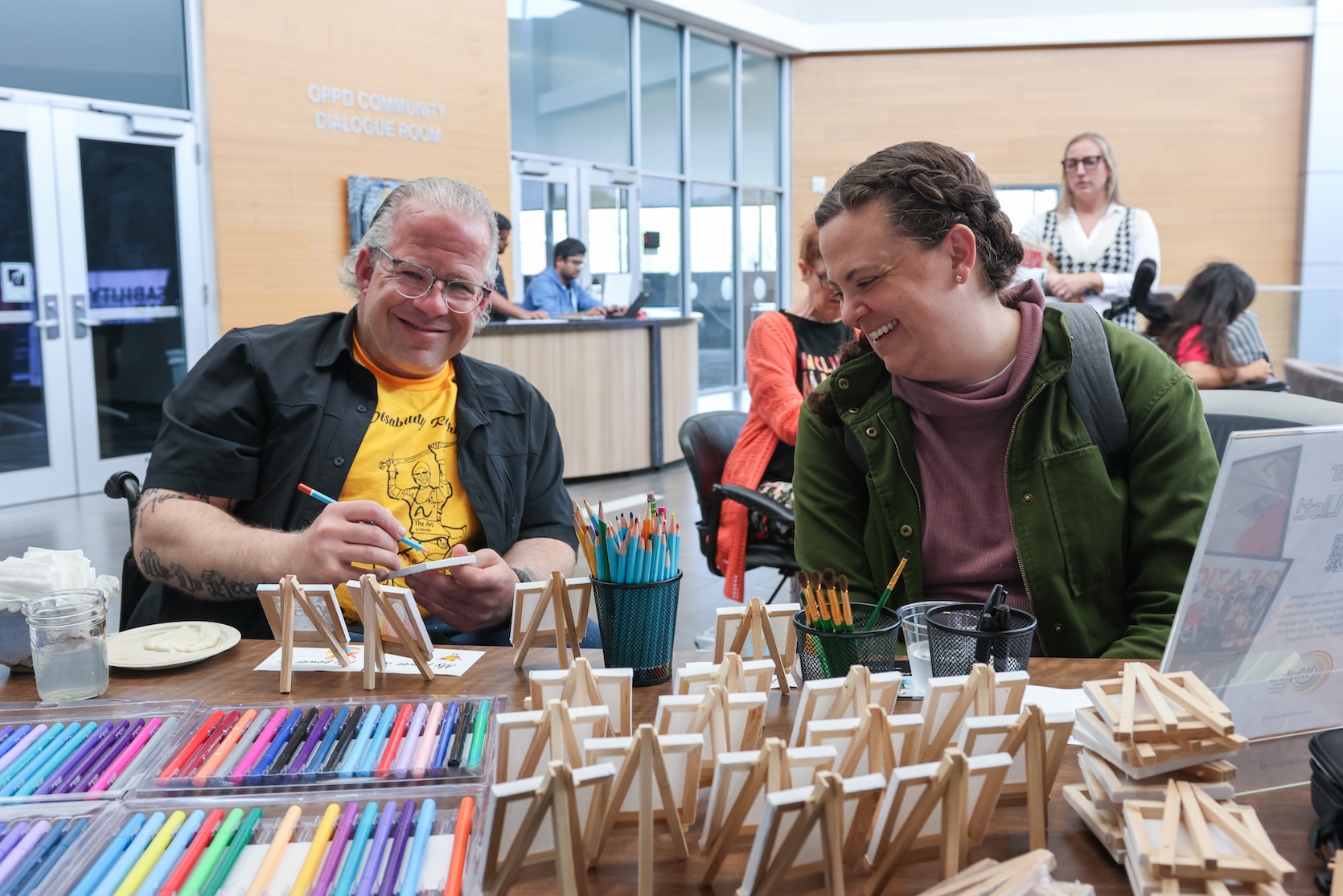 A white man with white hair slicked back. He is wearing glasses, a yellow t-shirt with the word “disability” on it, and a short sleeve black button up left unbuttoned. Beside him is a white woman with her brown hair styled back. She is wearing a red-purple top with a green jacket over it. They are sitting at a craft table.