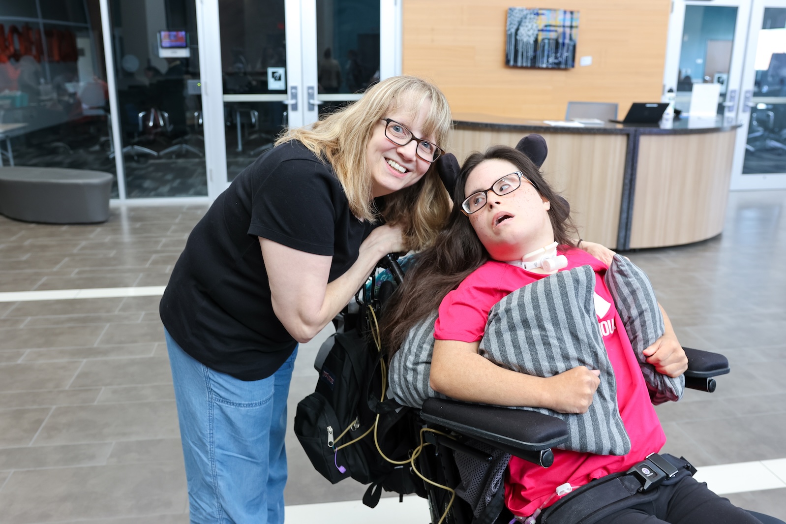 A white woman with long blonde hair with bangs, wearing glasses, a black t-shirt, and denim jeans smiles and poses with a young white woman with long brown hair. She is wearing glasses, a pink t-shirt, and black jeans. She is sitting in a wheelchair.