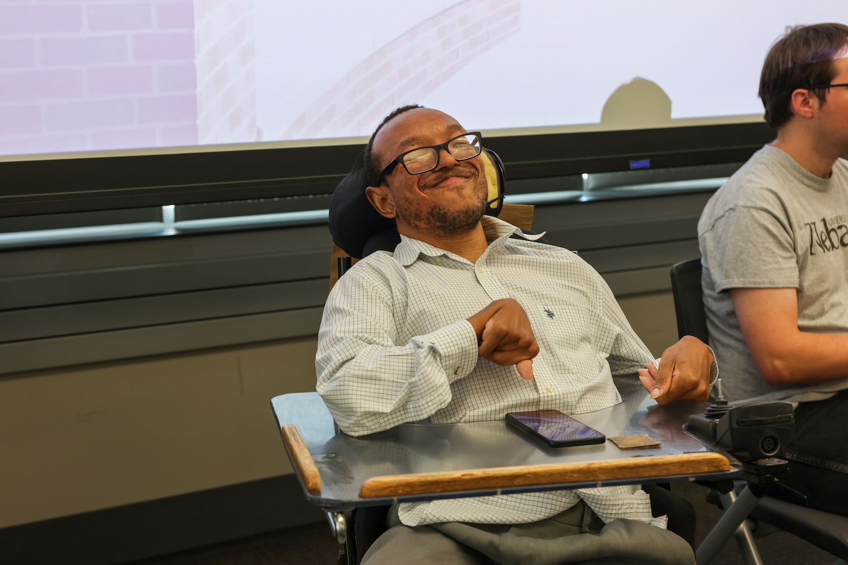 A Black man with glasses, wearing a white gingham button up, sits in his powerchair, smiling.