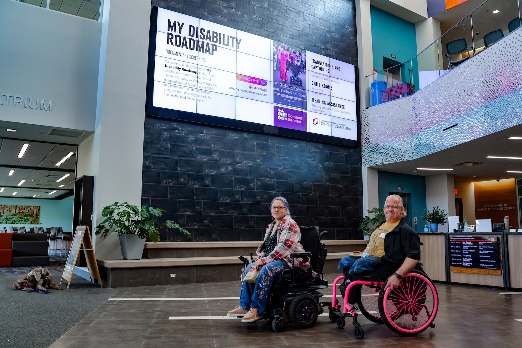 A white woman and white man, both in wheelchairs, smile to the camera. Above them is an electronic sign that says My Disability Roadmap.