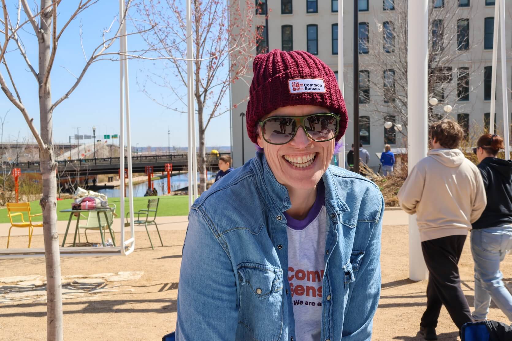A white woman wearing a red beanie, sunglasses, a Common Senses shirt, and denim jacket smiling. She is standing outside. The photo is a closeup. The Riverfront is in the background.