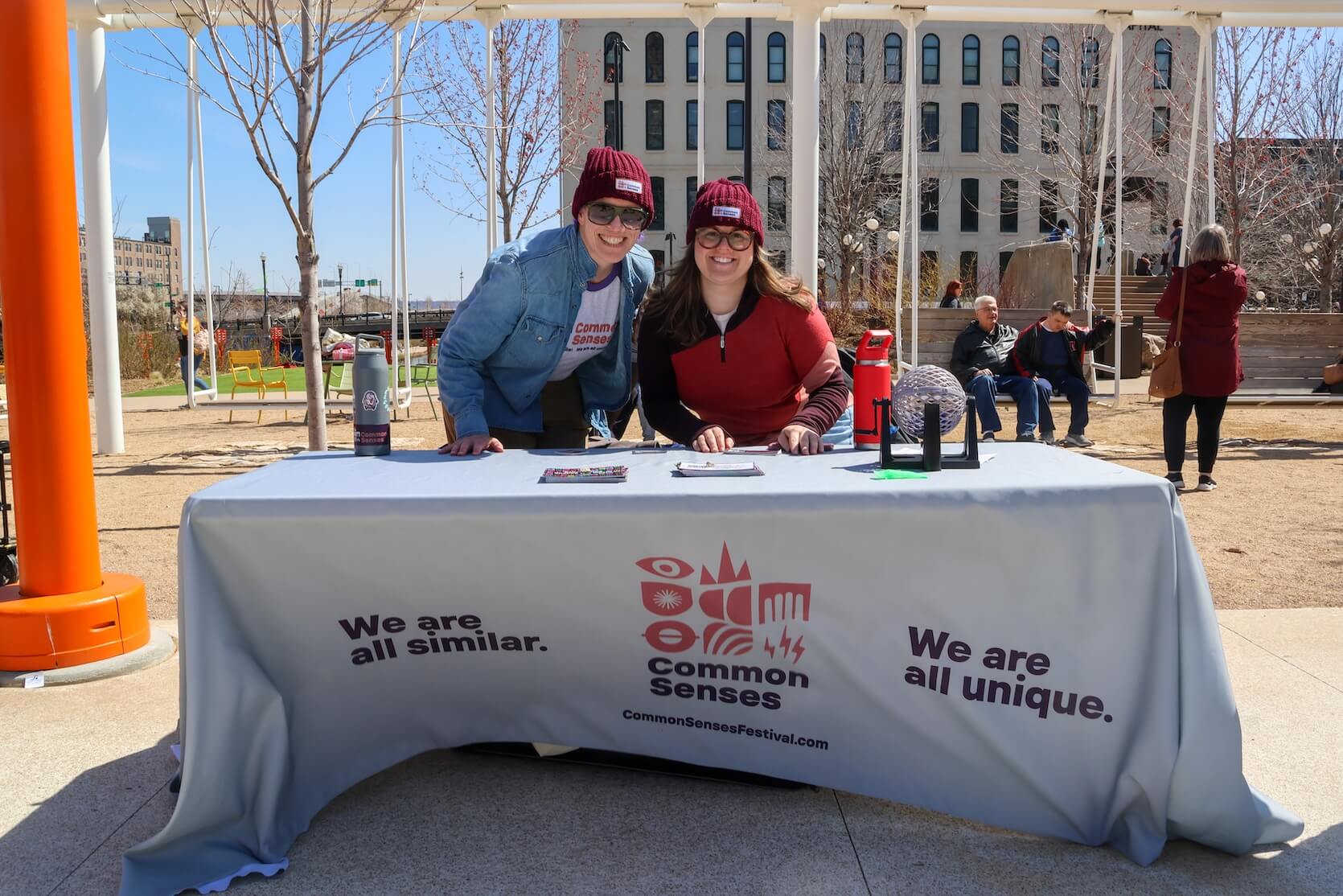 Two white women outside, standing at a table, smiling. They are in cold weather attire. Common Senses Festival marketing material laid out. A table cloth promoting their organization and event covers the table. People are sitting on the swings behind them.