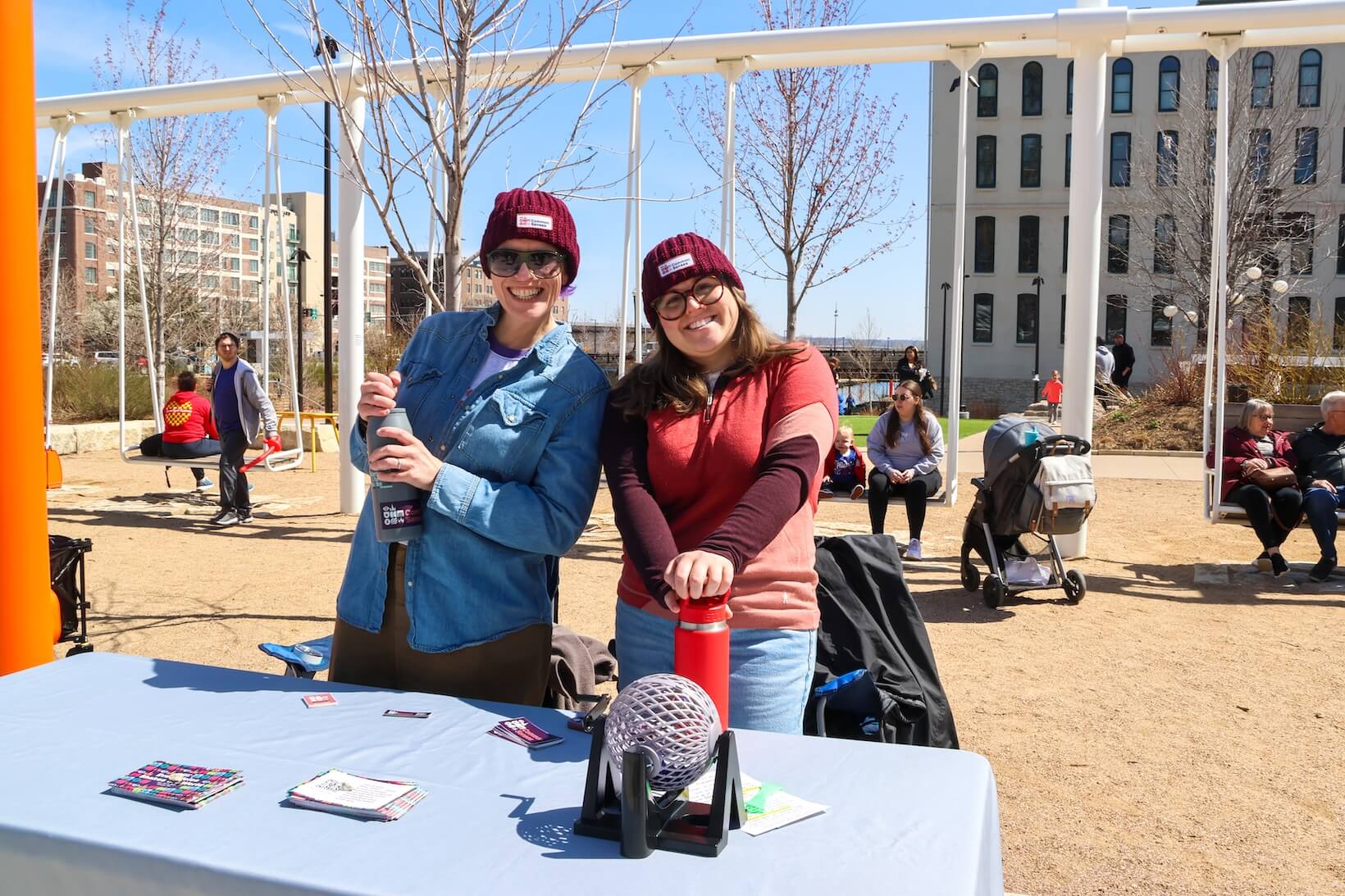 Two white women outside, standing at a table, smiling. They are in cold weather attire. Common Senses Festival marketing material laid out. People are sitting on the swings behind them.