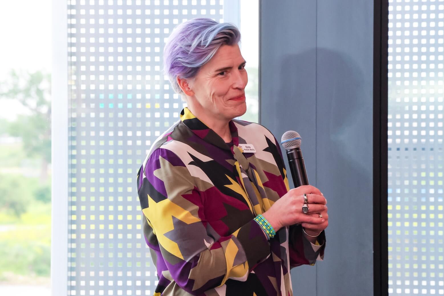 A white woman with short blue and purple hair, smiling while holding a microphone. She is wearing a multicolored abstract top.
