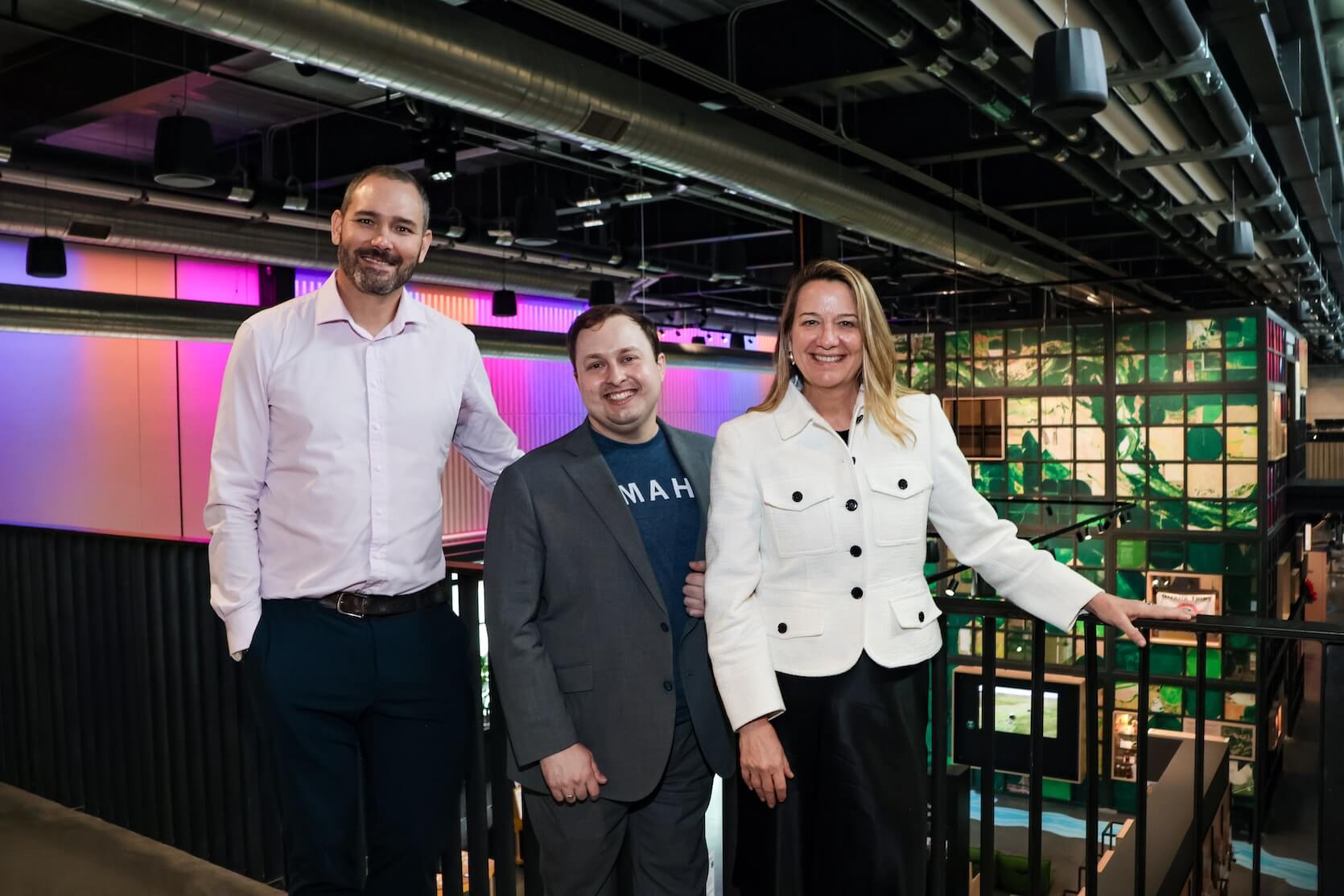 A tall man with short shaved hair and a beard, wearing a light purple button up shirt and denim jeans. A shorter man with short brown hair, a blue t-shirt that says “Omaha” in white letters, covered by a grey blazer and pants. The last woman has long blonde hair, and is wearing a white jacket and long black skirt.