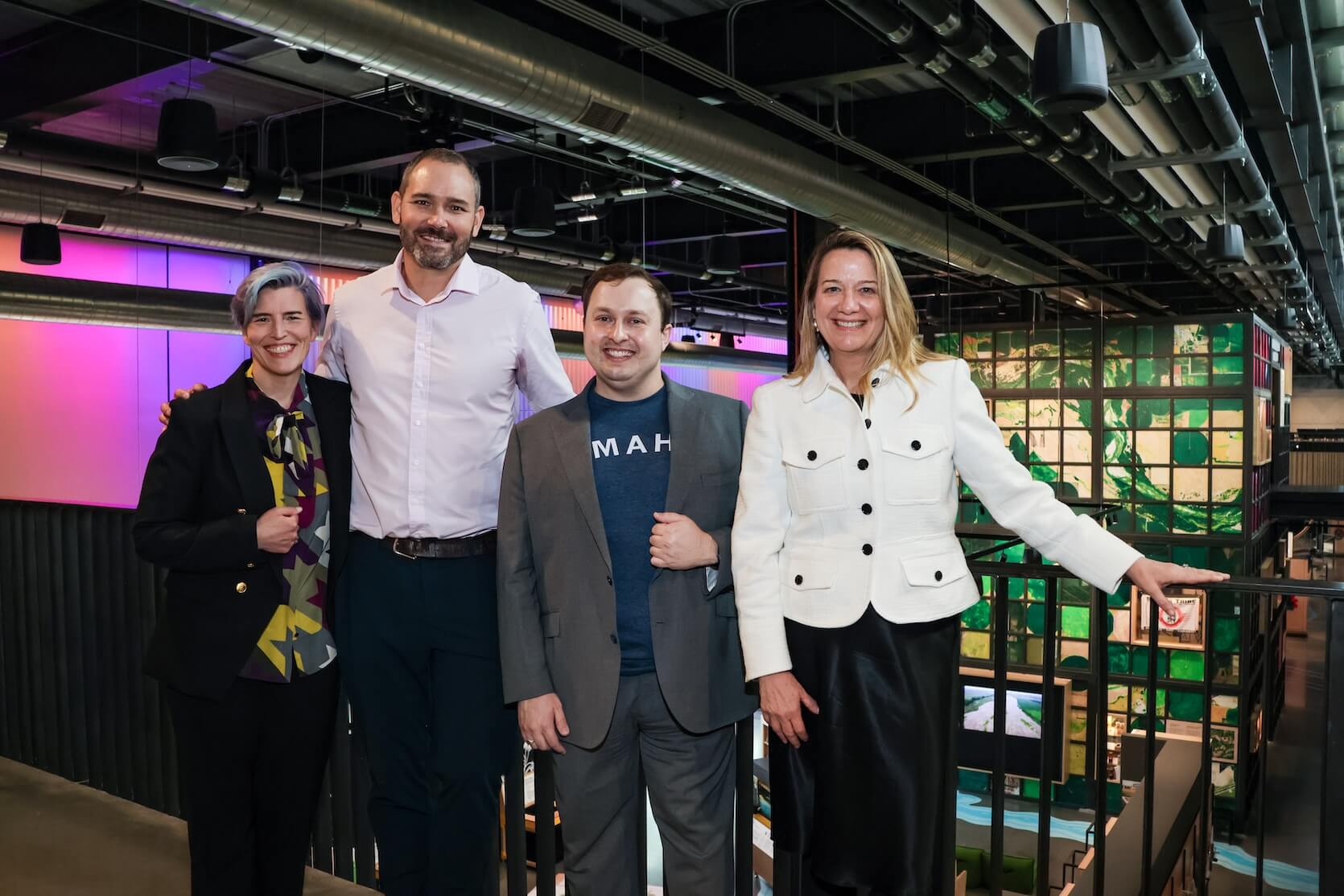 Two white women and two white men stand together, smiling. One woman with short blue and purple hair, wearing a multicolored abstract top, covered by a black blazer and pants. A tall man with short shaved hair and a beard, wearing a light purple button up shirt and denim jeans. A shorter man with short brown hair, a blue t-shirt that says “Omaha” in white letters, covered by a grey blazer and pants. The last woman has long blonde hair, and is wearing a white jacket and long black skirt.