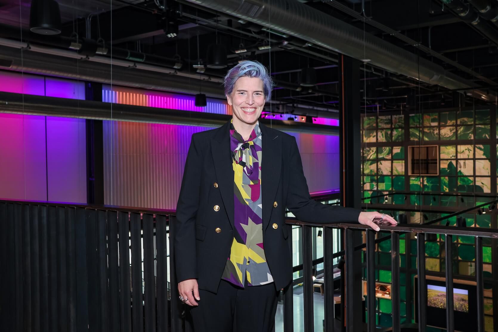 A white woman with short blue and purple hair, smiling. She is leaning against a railing. She is wearing a multicolored abstract top, covered by a black blazer and pants.