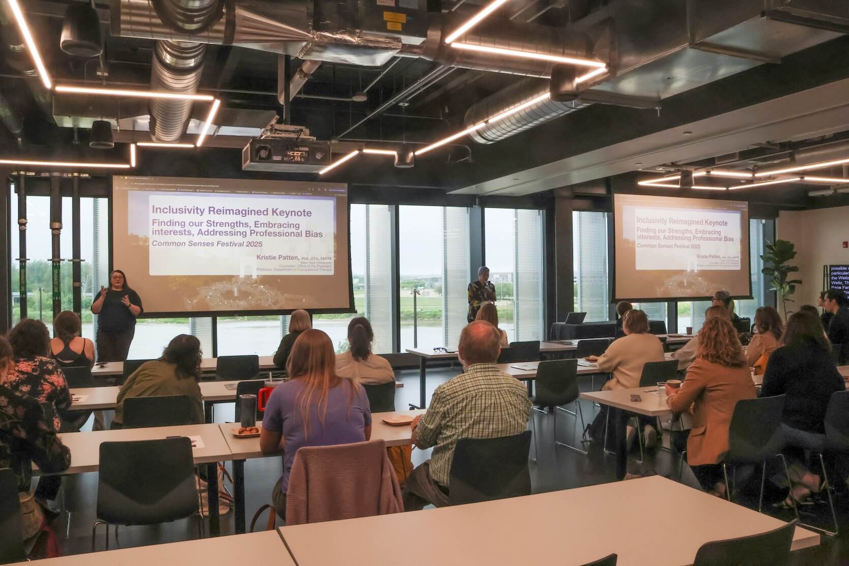 Back view of audience members in a conference room watching the speaker at the podium. An ASL interpreter is present, as well as Live Captions at the far right.