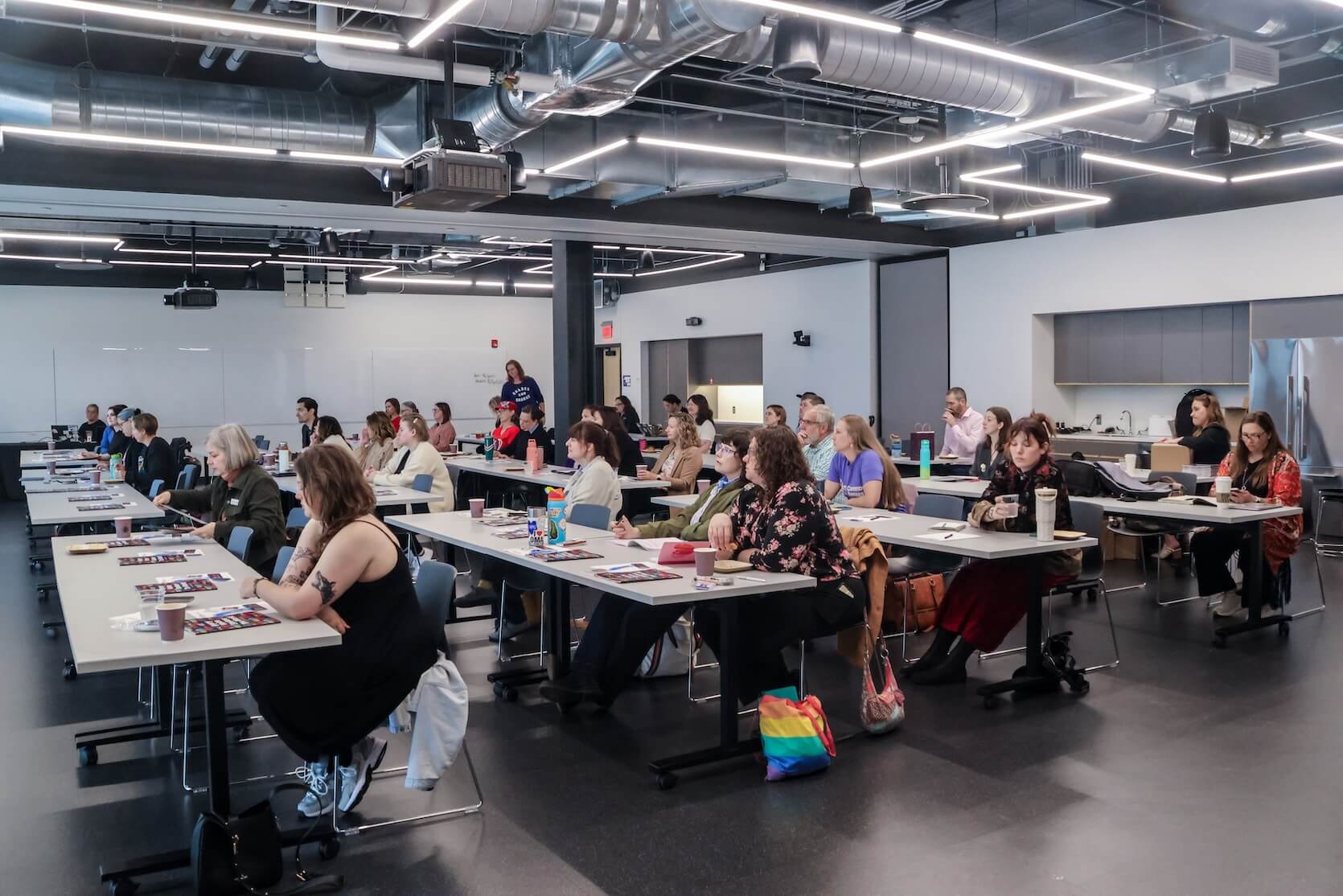 Many people sit as an audience at tables in a conference room. Some looking at the materials on the table, some looking at the speaker.