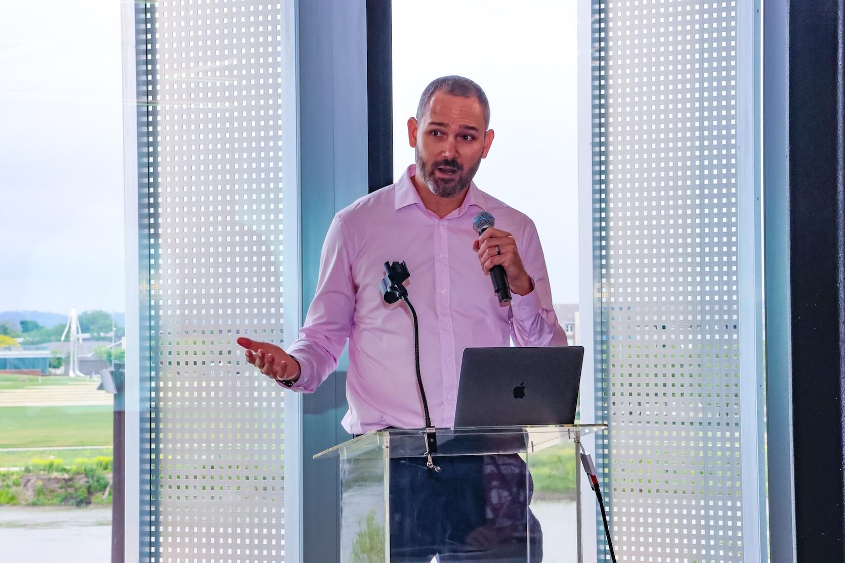 A white man with short shaved hair and a beard, wearing a light purple button up shirt and denim jeans, stands at a podium. He is holding a microphone and speaking. His laptop is open in front of him.