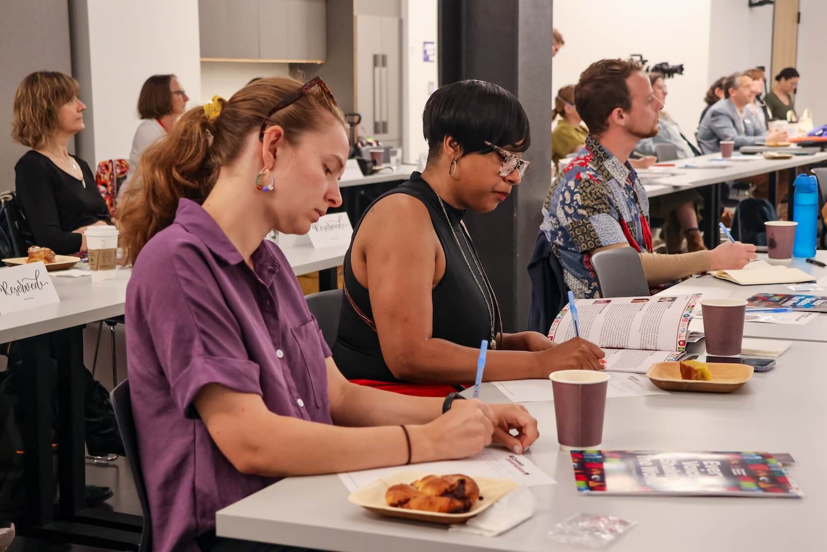Audience members sit, focused on the materials at their table. They are writing something down on paper. Other members are focused on the speaker.