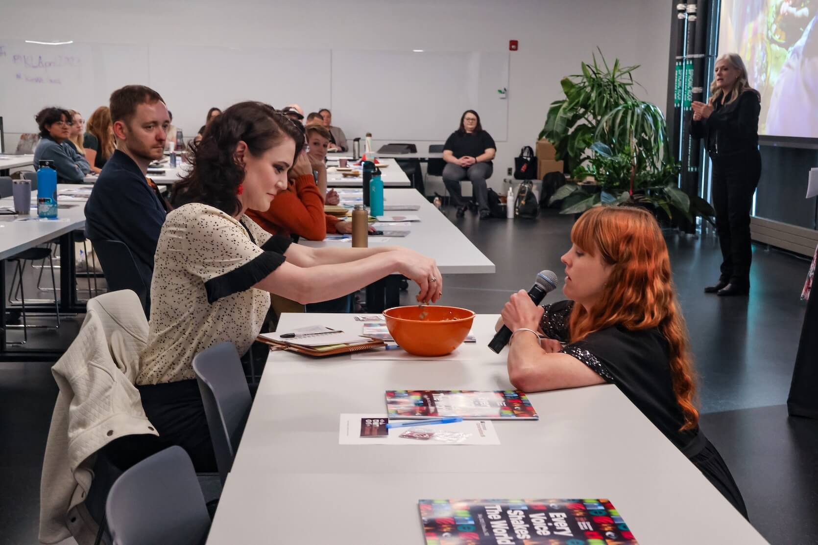 The speaker, a white woman with long red hair with bangs, squats down in front of an audience member, a white woman with pinned back curly shoulder length black hair. The audience member has her hands in an orange bowl. Other audience members are watching.